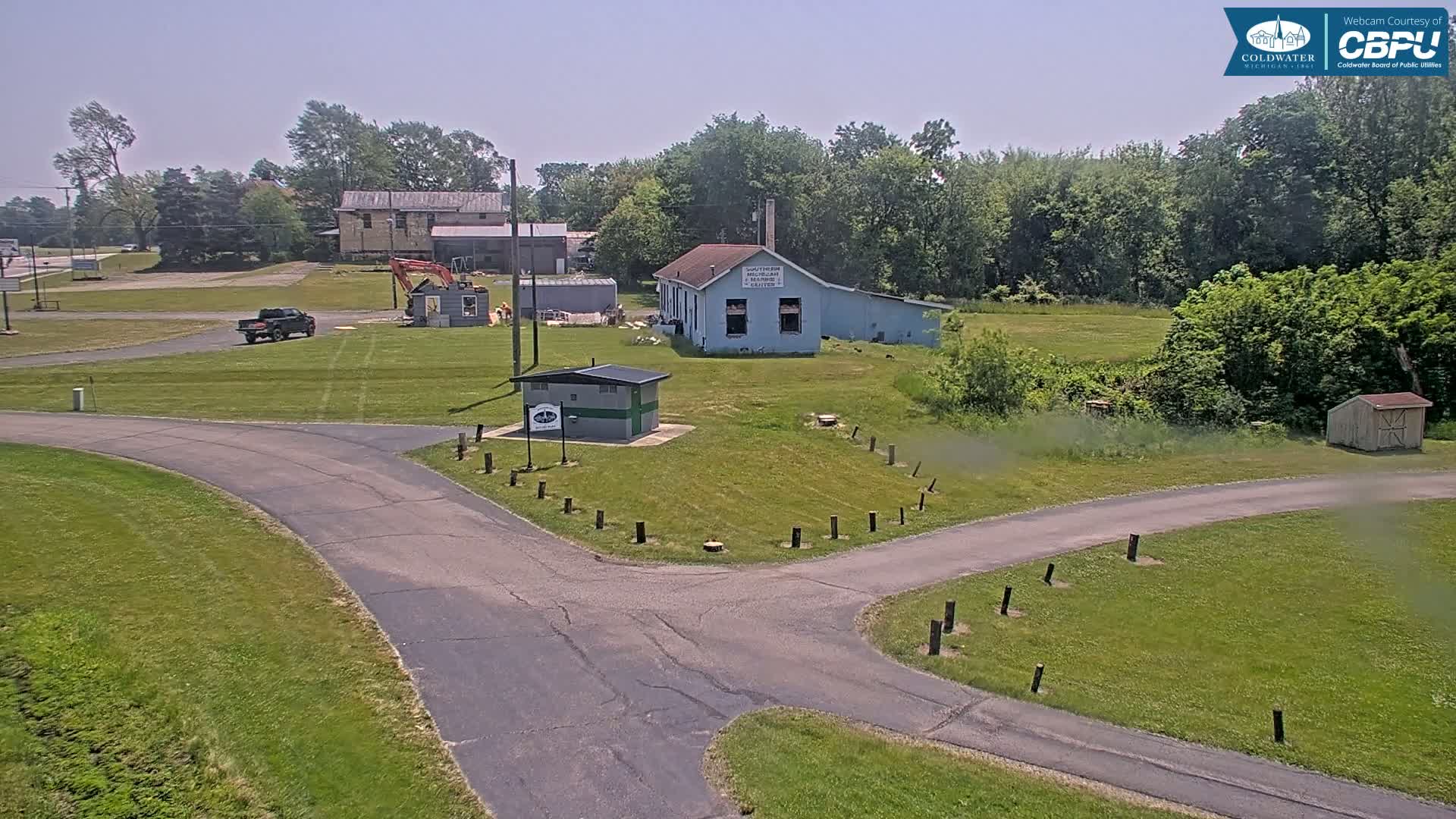 A paved road forks in a grassy area near a small blue building and a pickup truck, under a clear sky.