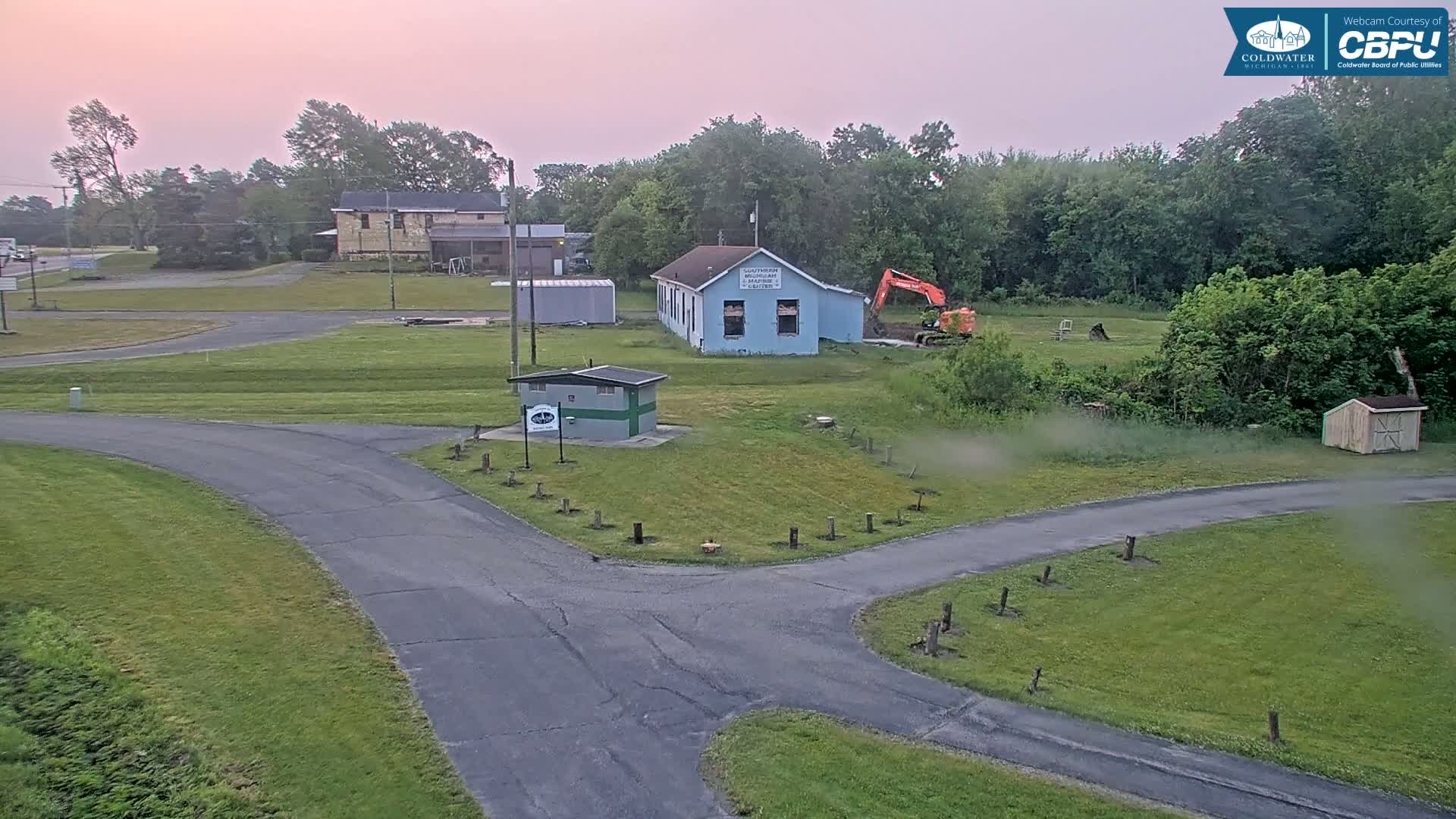 An aerial view shows a small town under a pink dawn sky, with a road intersecting near a small building and an excavator working near a larger building.
