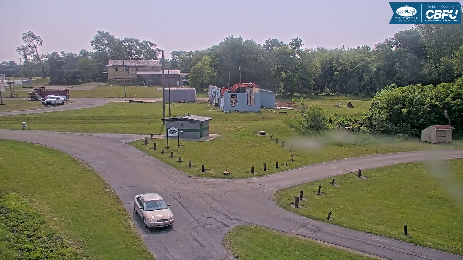 A car drives past a small building and a partially demolished structure on a sunny day in a grassy area with several trucks parked nearby.