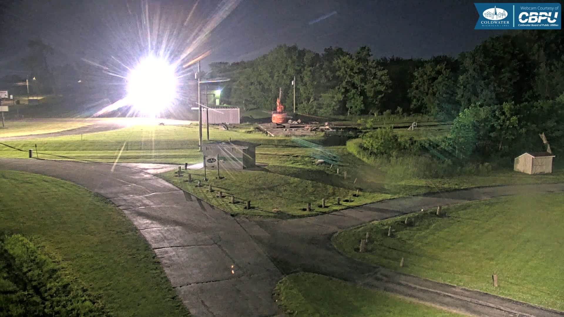A brightly lit paved area with grassy areas and a small building is visible at night, near a wooded area with an orange piece of construction equipment.