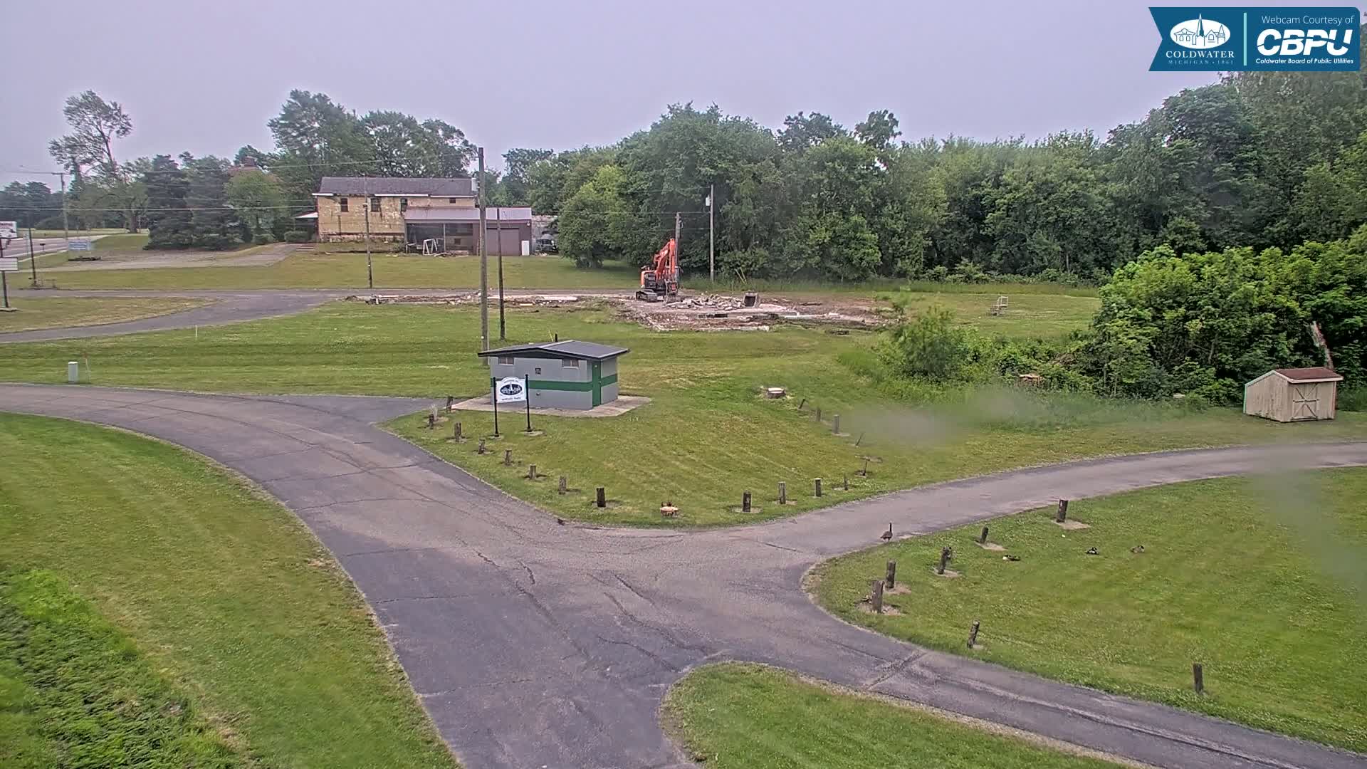 An overcast day reveals an excavator demolishing a structure in a grassy area near a road and small buildings, with several birds near a paved intersection.