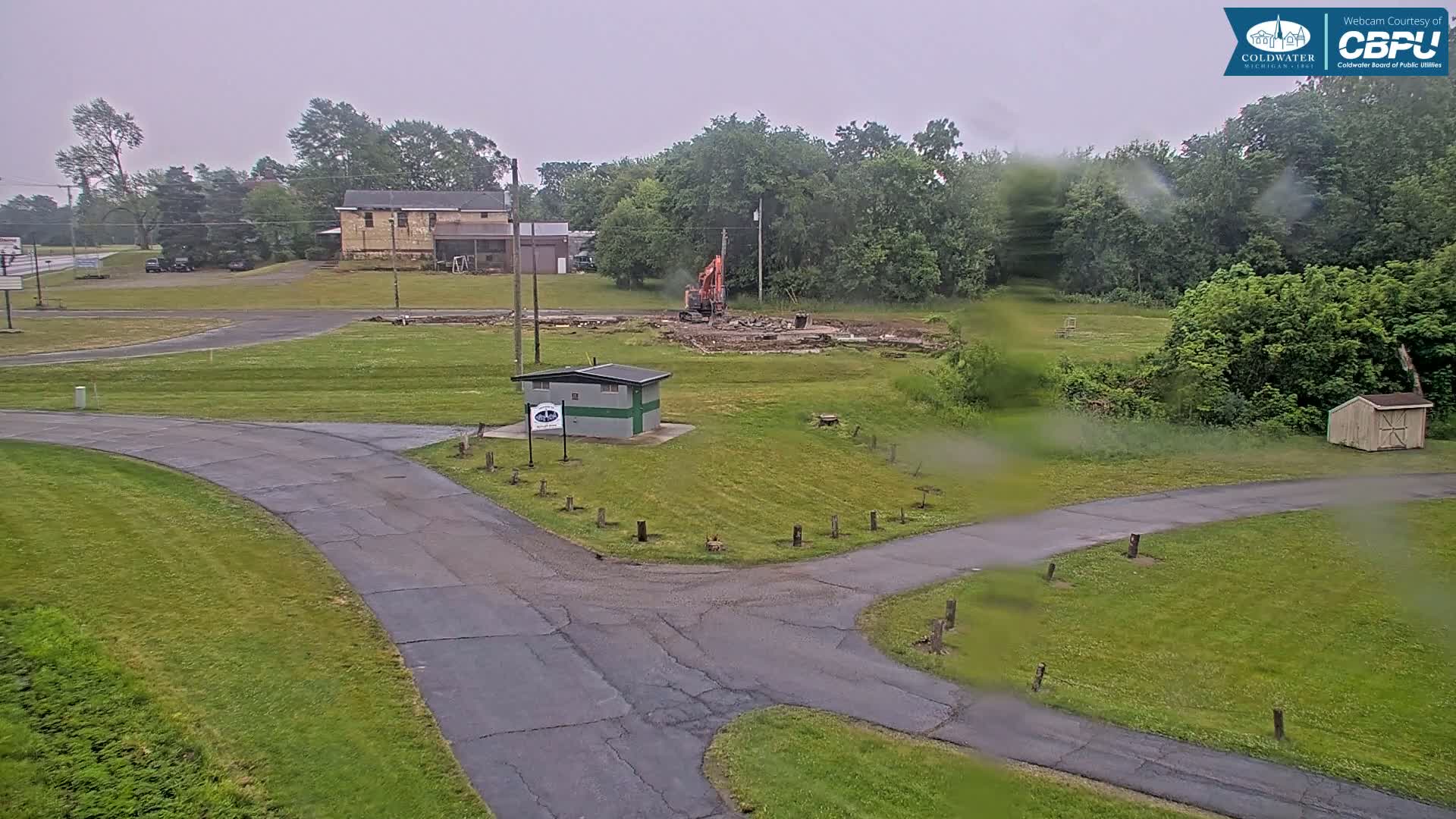 An overcast day shows an excavator demolishing a structure in a grassy area near a roadway and small building, with trees and other buildings in the background.