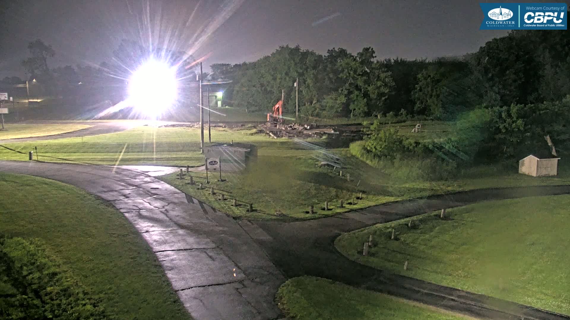 A brightly lit construction site with an excavator is visible at night under overcast skies, near a roadway and grassy areas.