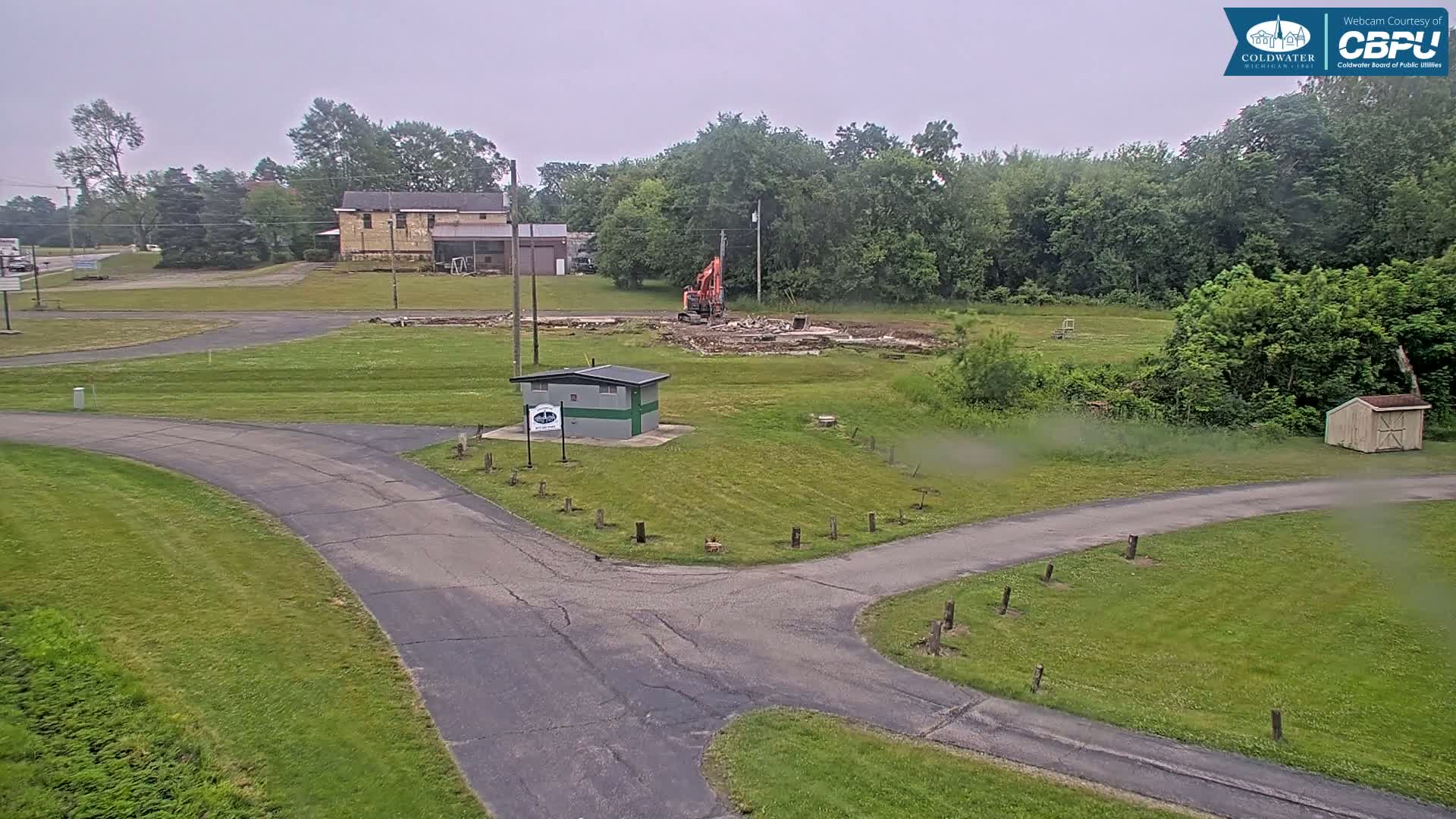 A small utility building sits near a paved road intersection in a grassy area where a demolition is taking place under an overcast sky.