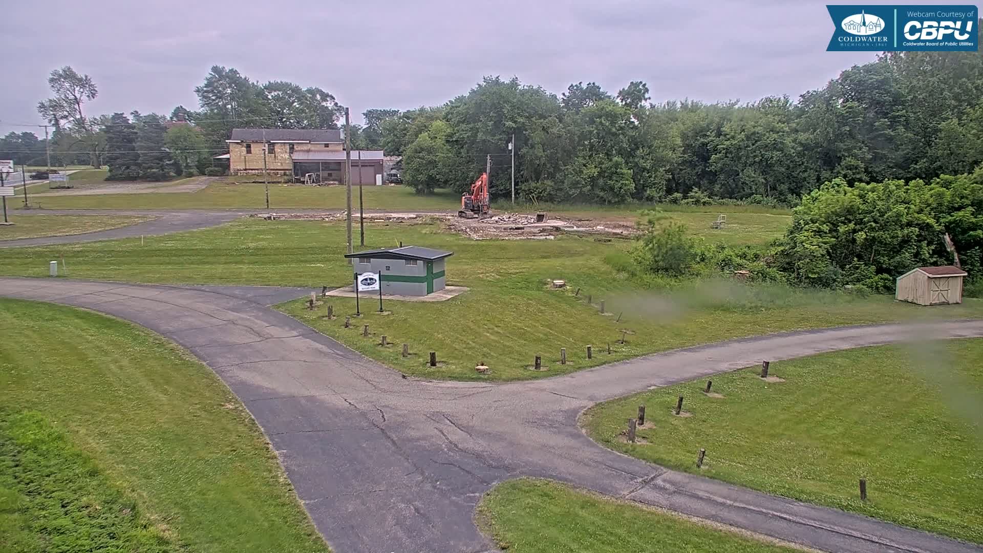 An overcast day shows an excavator demolishing a structure in a grassy area with paved roads and a small building nearby, surrounded by trees.