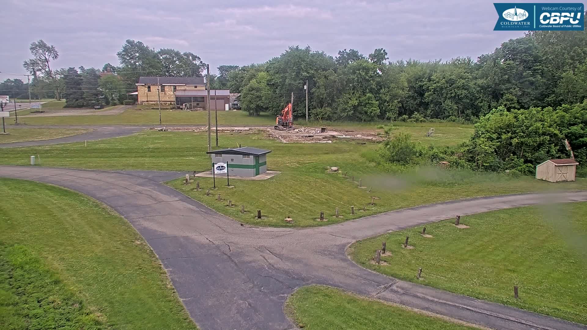 An overcast day shows an excavator demolishing a structure in a grassy area with paved roads and a small building nearby, surrounded by trees.