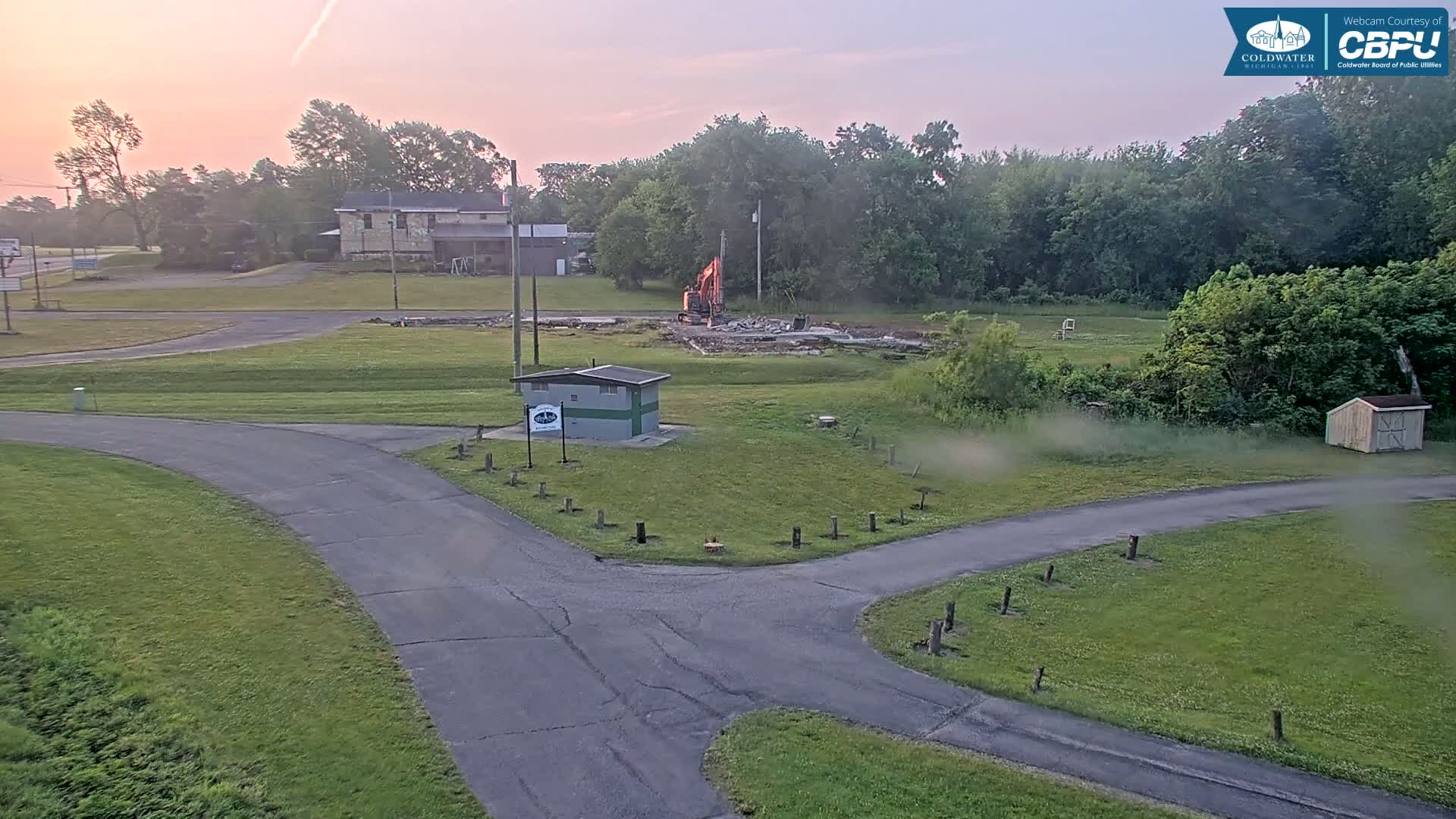 A demolition crew uses a backhoe to tear down a building in a grassy area under a pale sunrise sky.