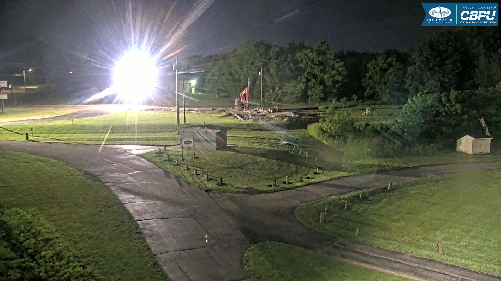 A brightly lit construction site at night, featuring a small building, an excavator, and intersecting roadways surrounded by grassy areas and trees.