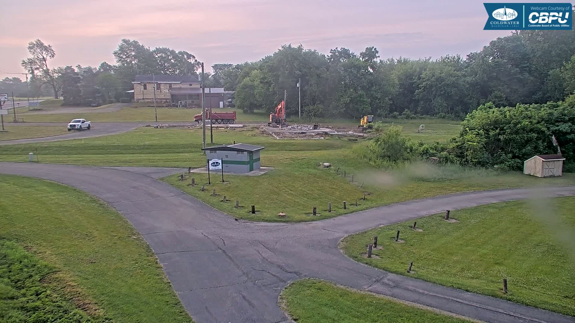 A demolition site with two excavators is visible next to a small building and a roadway under a dawn sky.