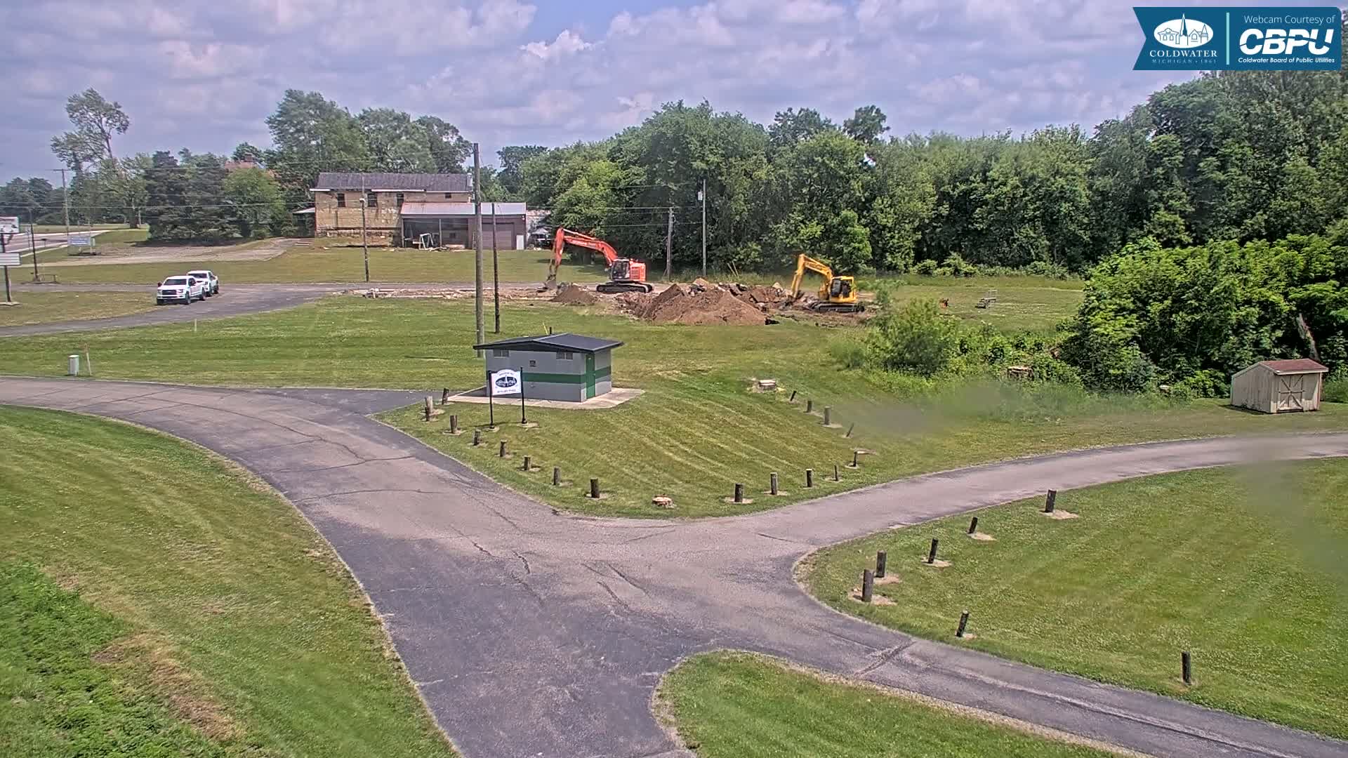 Two excavators are digging up earth near a small building in a grassy area under a partly cloudy sky.