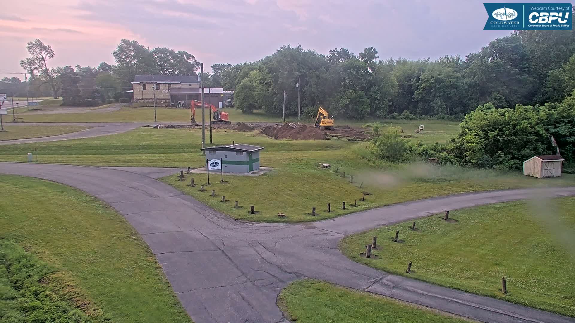 Two excavators are working on a construction site near a small building and a road, under an overcast sky.