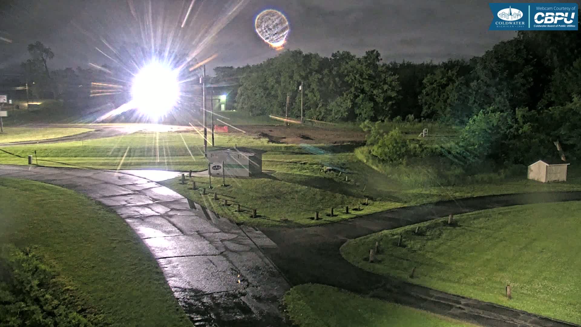 A nighttime view of a grassy area with roads and small buildings, under a cloudy sky with a bright light source creating lens flares.