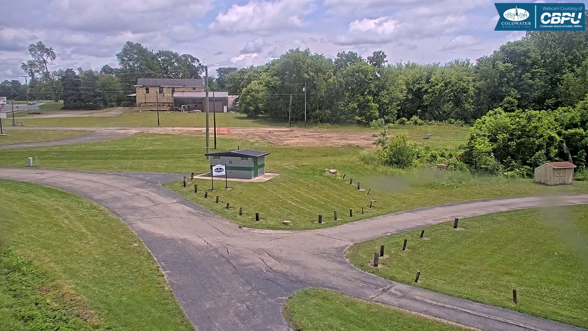 A paved path intersects in a grassy area with a small building, a larger building in the background, and a partly cloudy sky.