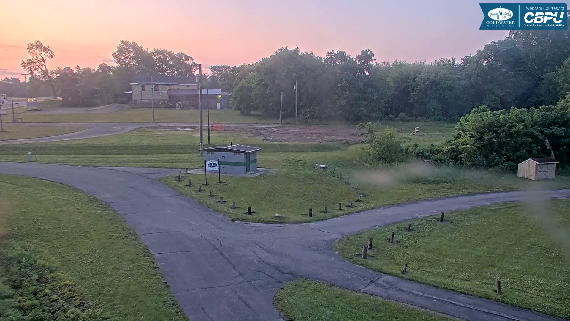 A paved roadway curves through a grassy area with a small building and trees, under a pink-tinged sunrise sky.