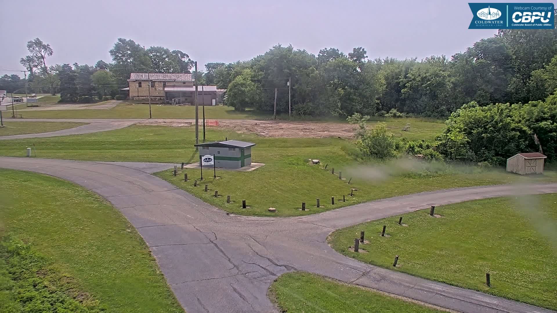 A paved road intersects with another, passing a small restroom building and a cleared area next to a wooded area under an overcast sky.