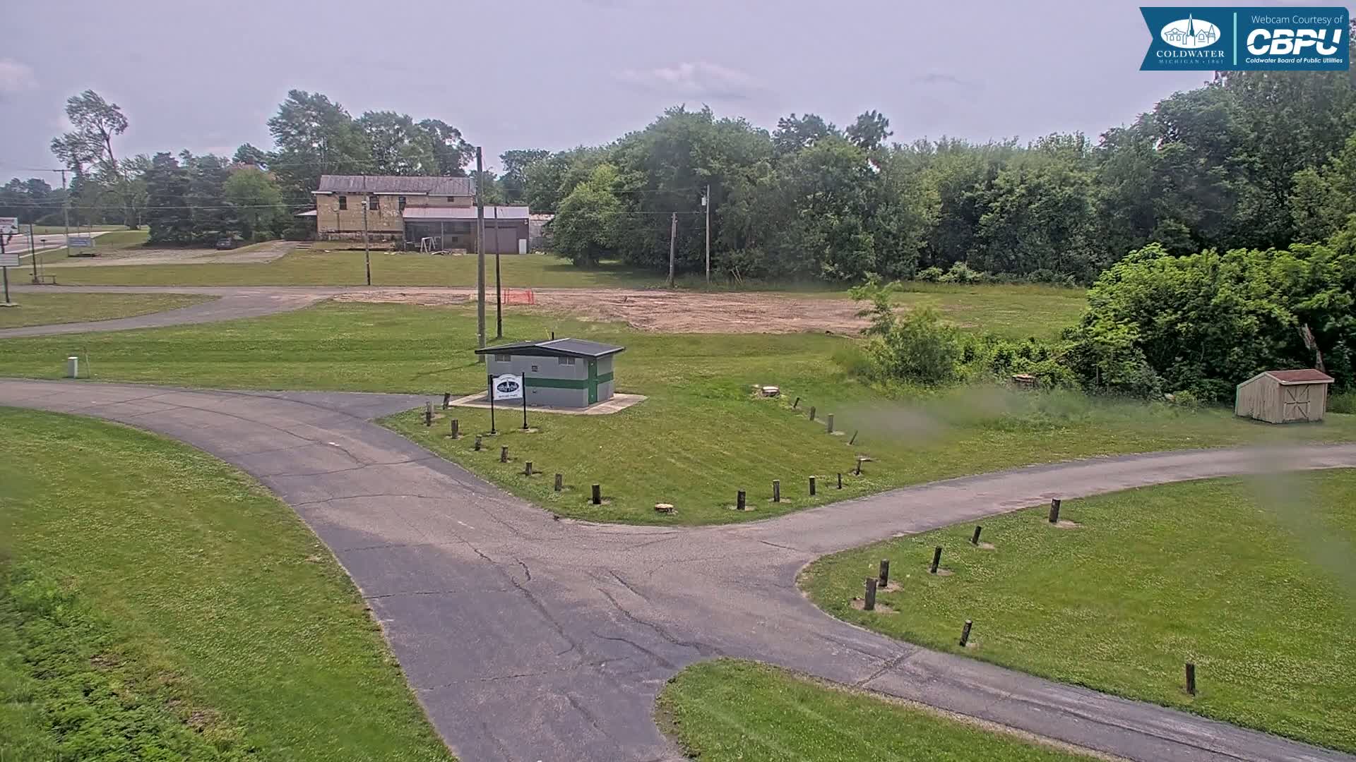 A paved pathway intersects in a grassy area with a small building and a larger building in the background under an overcast sky.