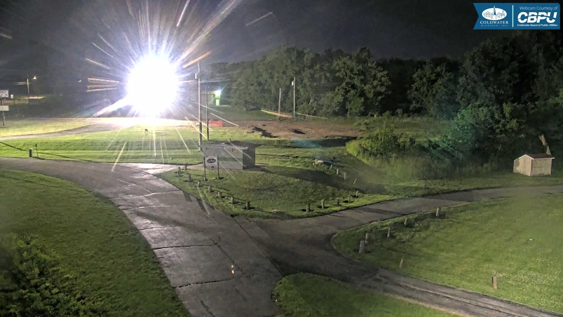 A brightly lit grassy area at night, featuring a small building, paved pathways, and a wooded backdrop.