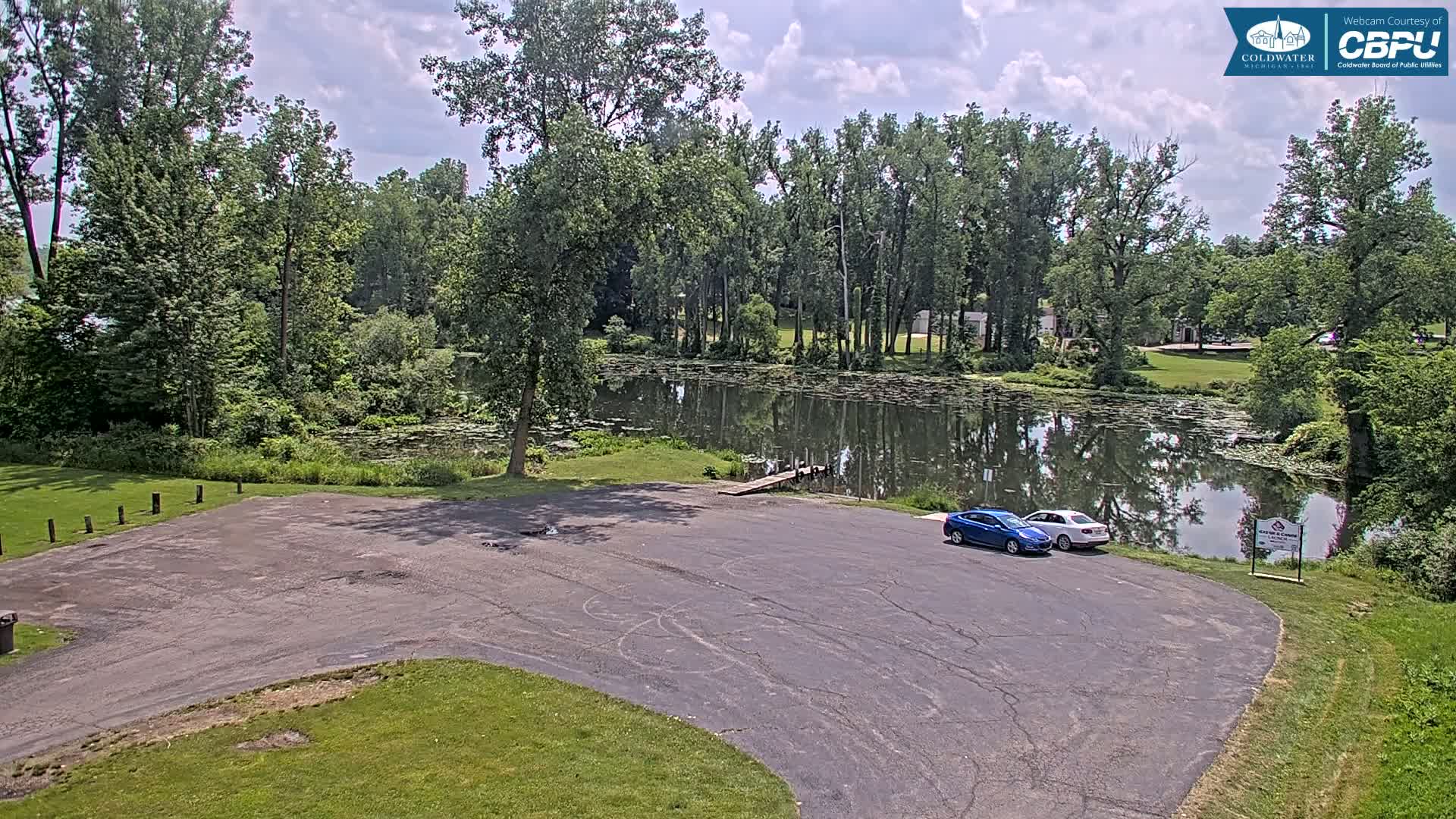 A partly cloudy day reveals a small, calm lake surrounded by lush green trees and a small, empty parking lot with two cars parked near a boat ramp.