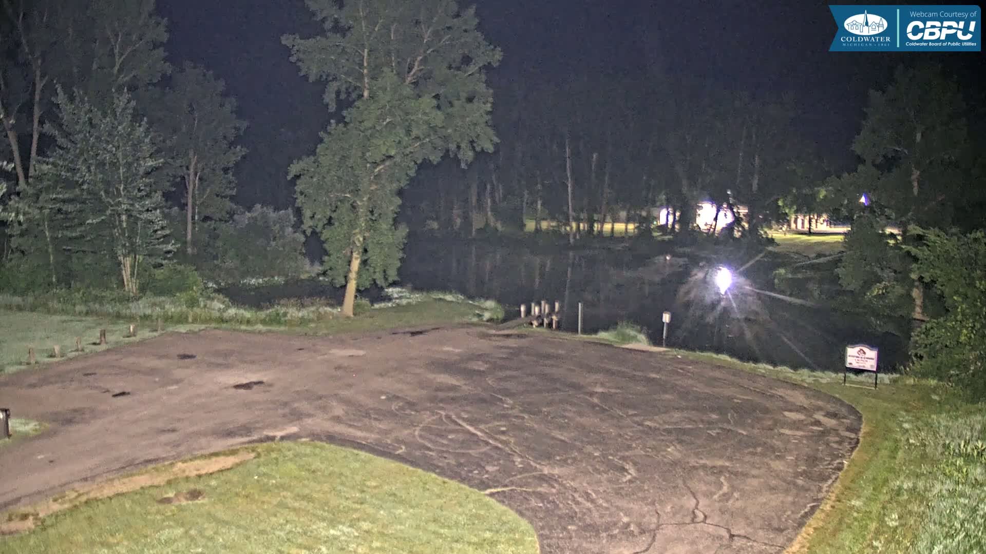 An empty, dark asphalt parking lot at night next to a calm body of water surrounded by trees.