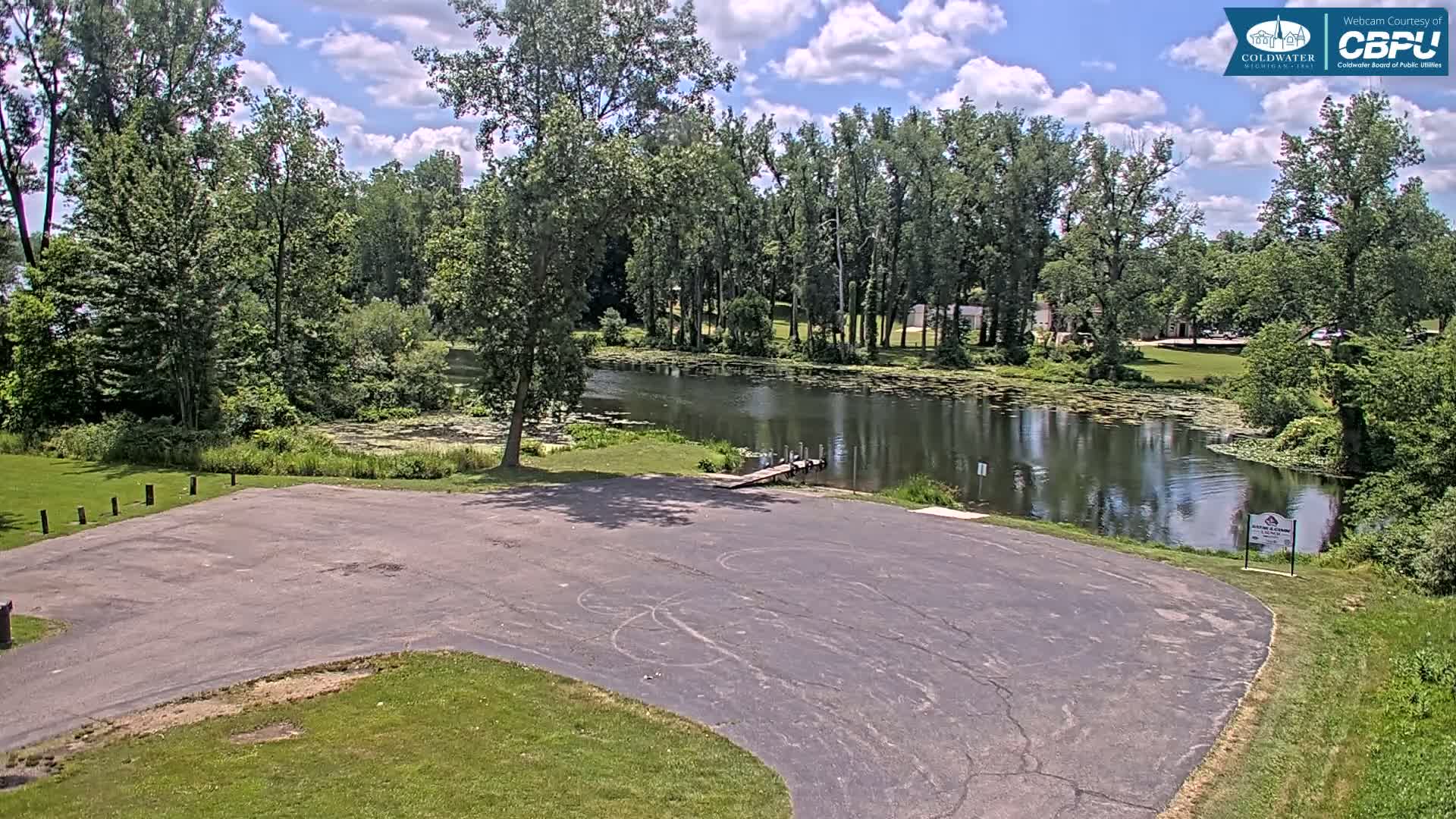 An empty asphalt parking lot overlooks a calm pond surrounded by lush green trees under a partly cloudy sky.