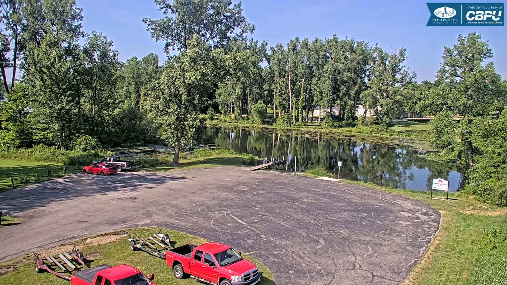 A calm, sunny day shows an empty boat launch parking lot with several red vehicles and boat trailers near a tranquil pond surrounded by lush green trees.
