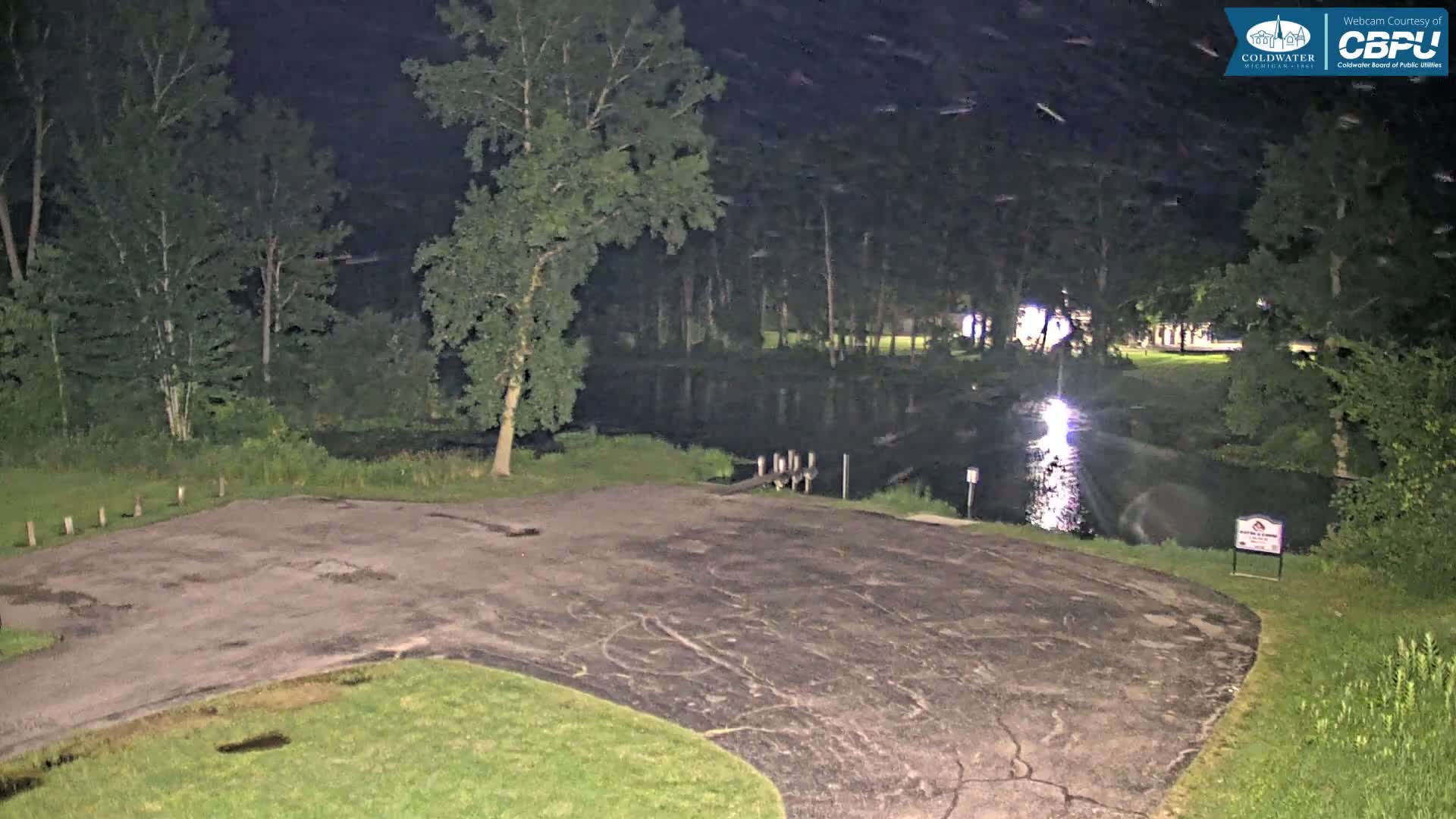 A dark, empty asphalt parking lot at night next to a calm, reflective body of water surrounded by trees.