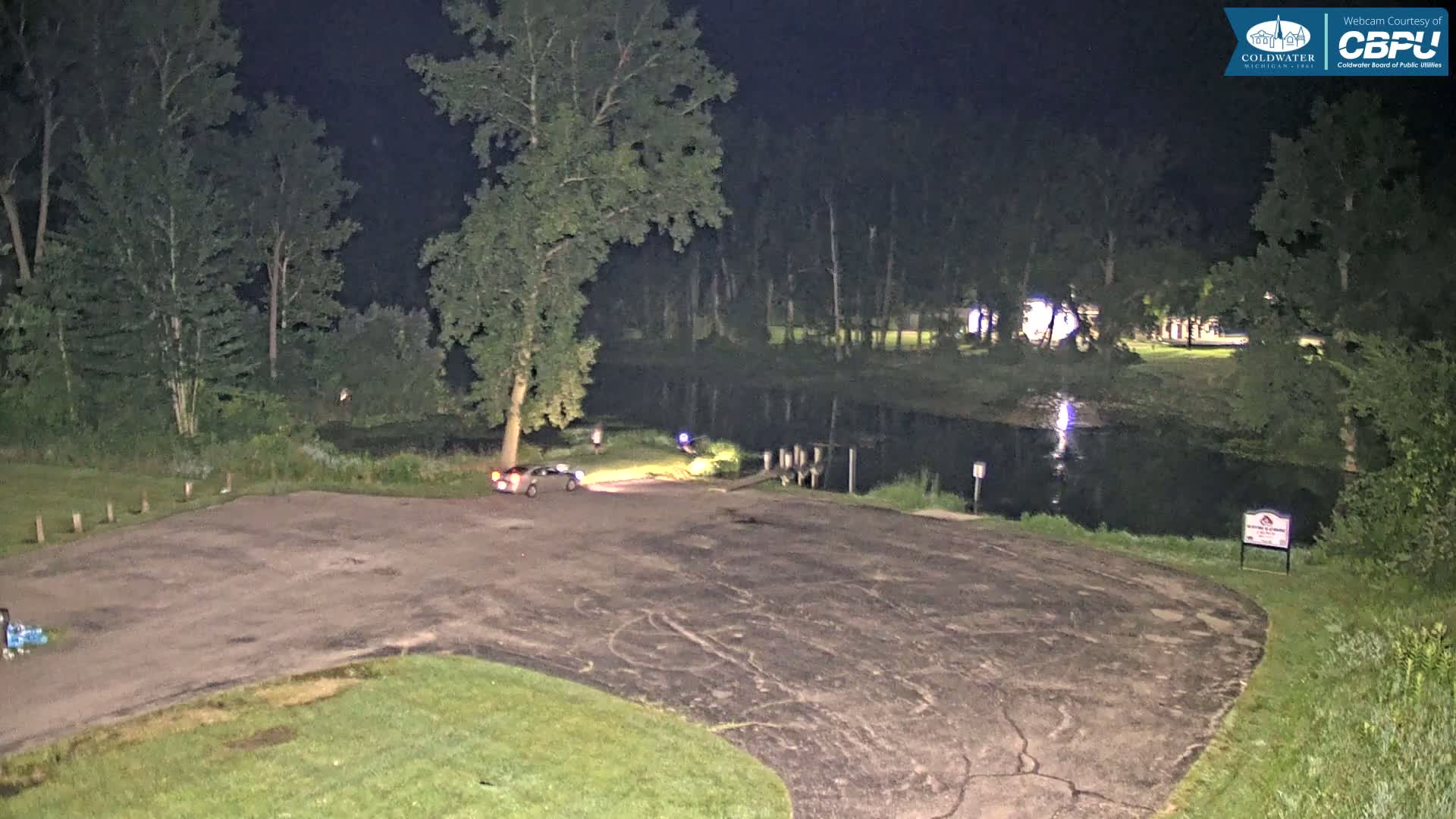 A dark, empty asphalt parking lot at night next to a calm, reflective body of water surrounded by trees.