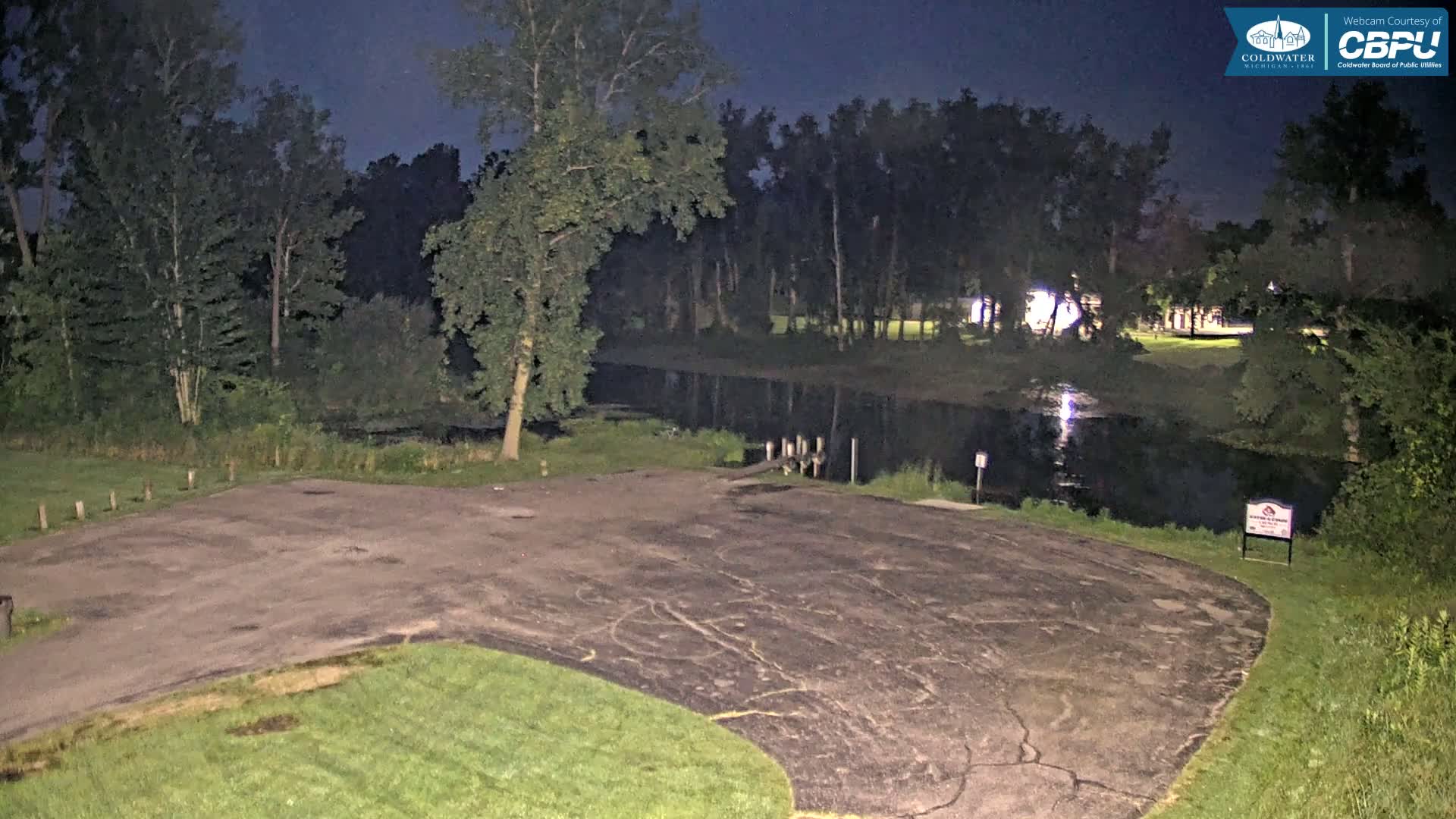 A dark asphalt boat launch area, bordered by grass and trees, sits next to a calm body of water under a night sky.