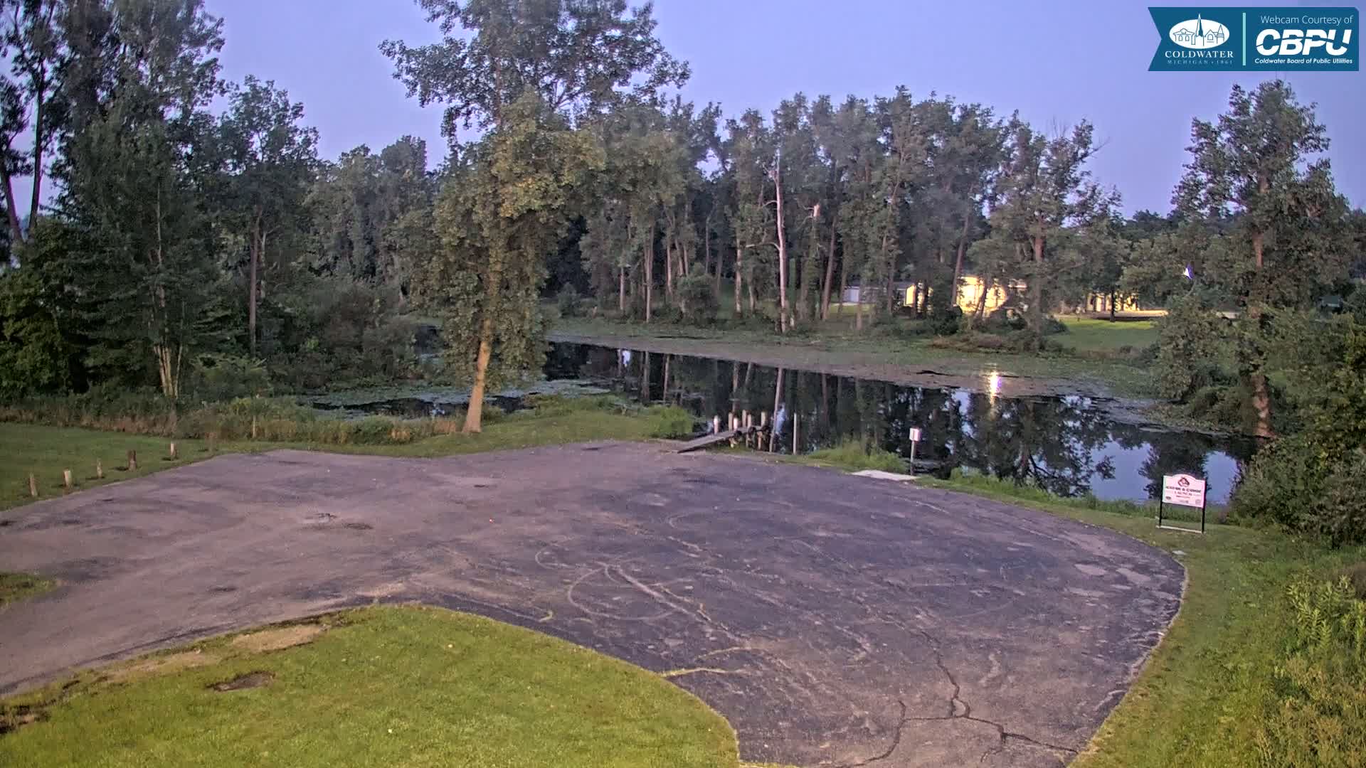 A dark asphalt boat launch area, bordered by grass and trees, sits next to a calm body of water under a night sky.