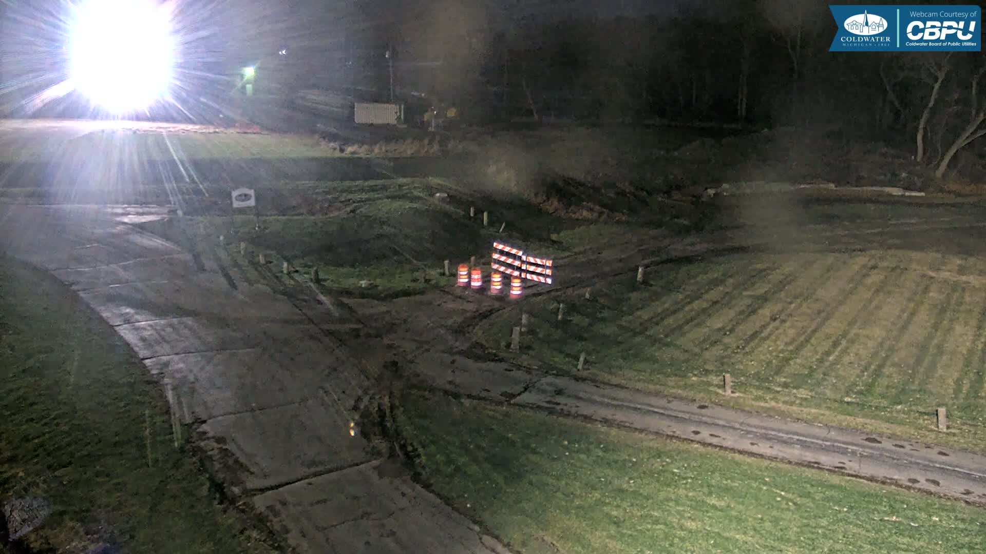 A bright light illuminates a damp, overcast outdoor scene at night, revealing paved and grassy paths with construction barriers and orange cones blocking a dirt road.