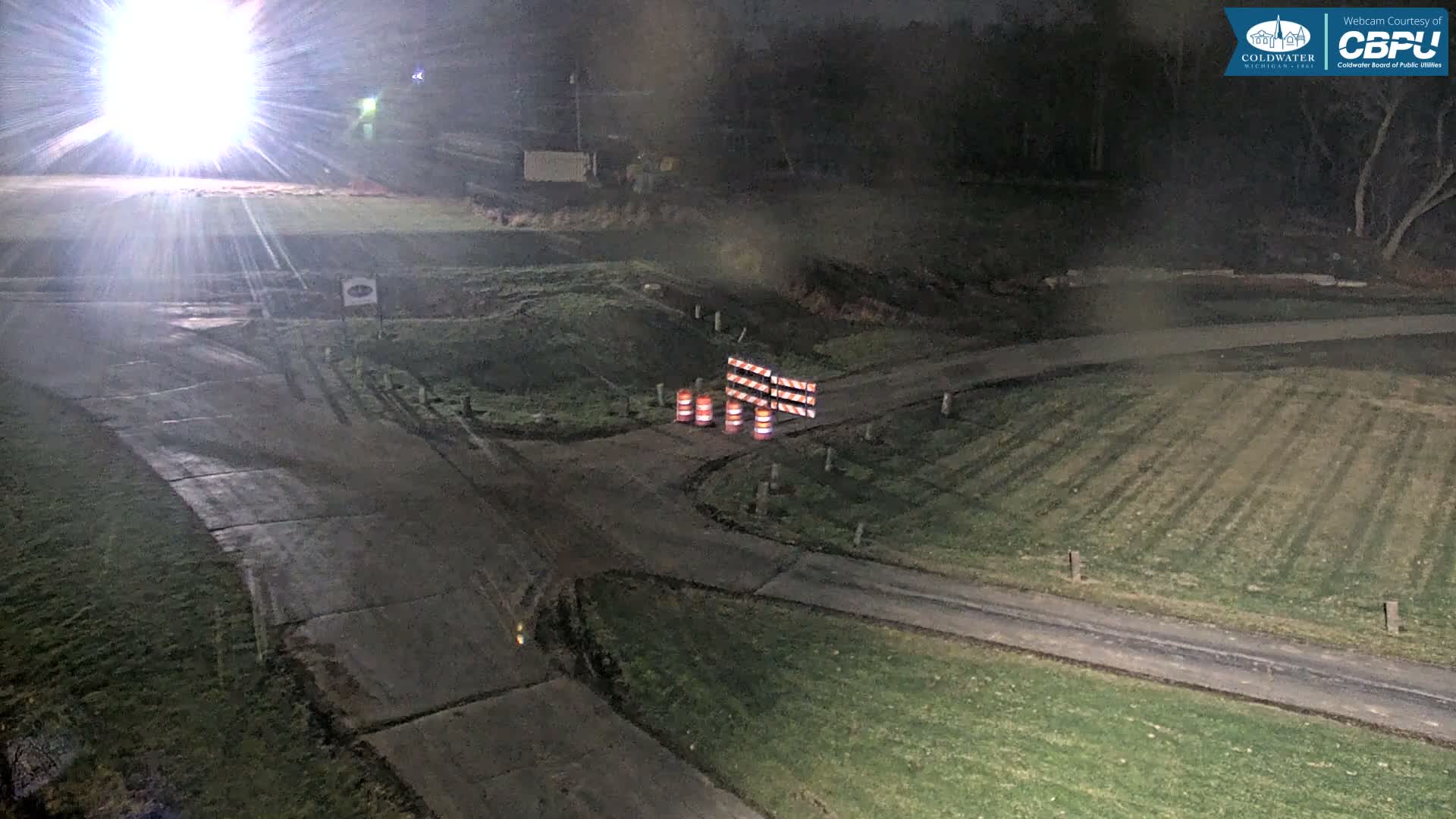 A misty night reveals a network of damp, unpaved paths and grassy terrain, brightly illuminated by a strong artificial light on the left, with striped traffic barrels and a barrier blocking a central intersection.