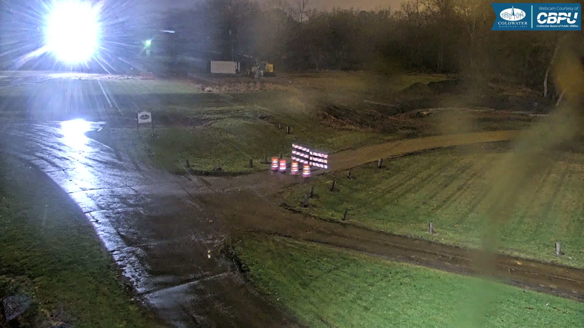 The image shows a rainy night scene outdoors, with wet asphalt and grassy terrain, featuring dirt paths blocked by construction barriers and cones under a bright light source.