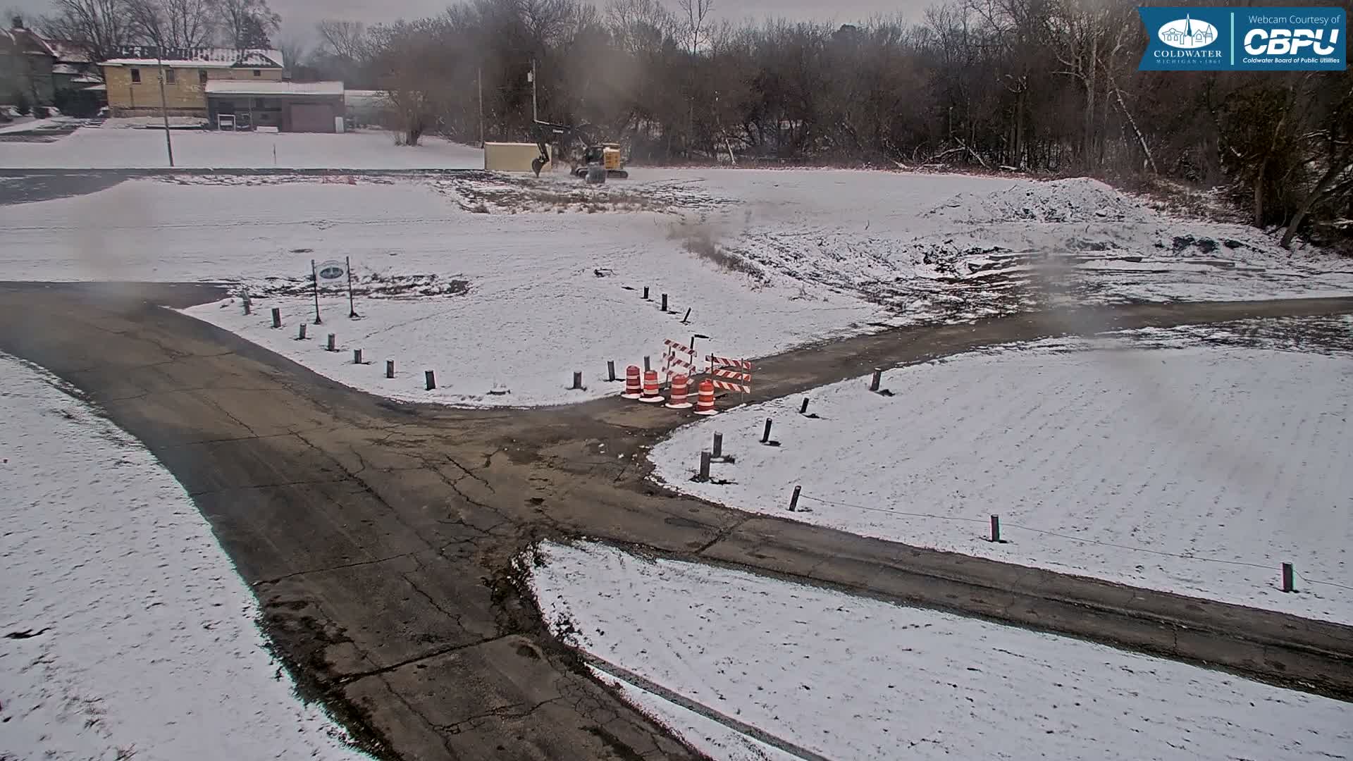 A winter scene shows a light covering of snow across fields and along cracked asphalt paths, where an excavator is working in the distance near traffic cones and barriers, under an overcast sky.