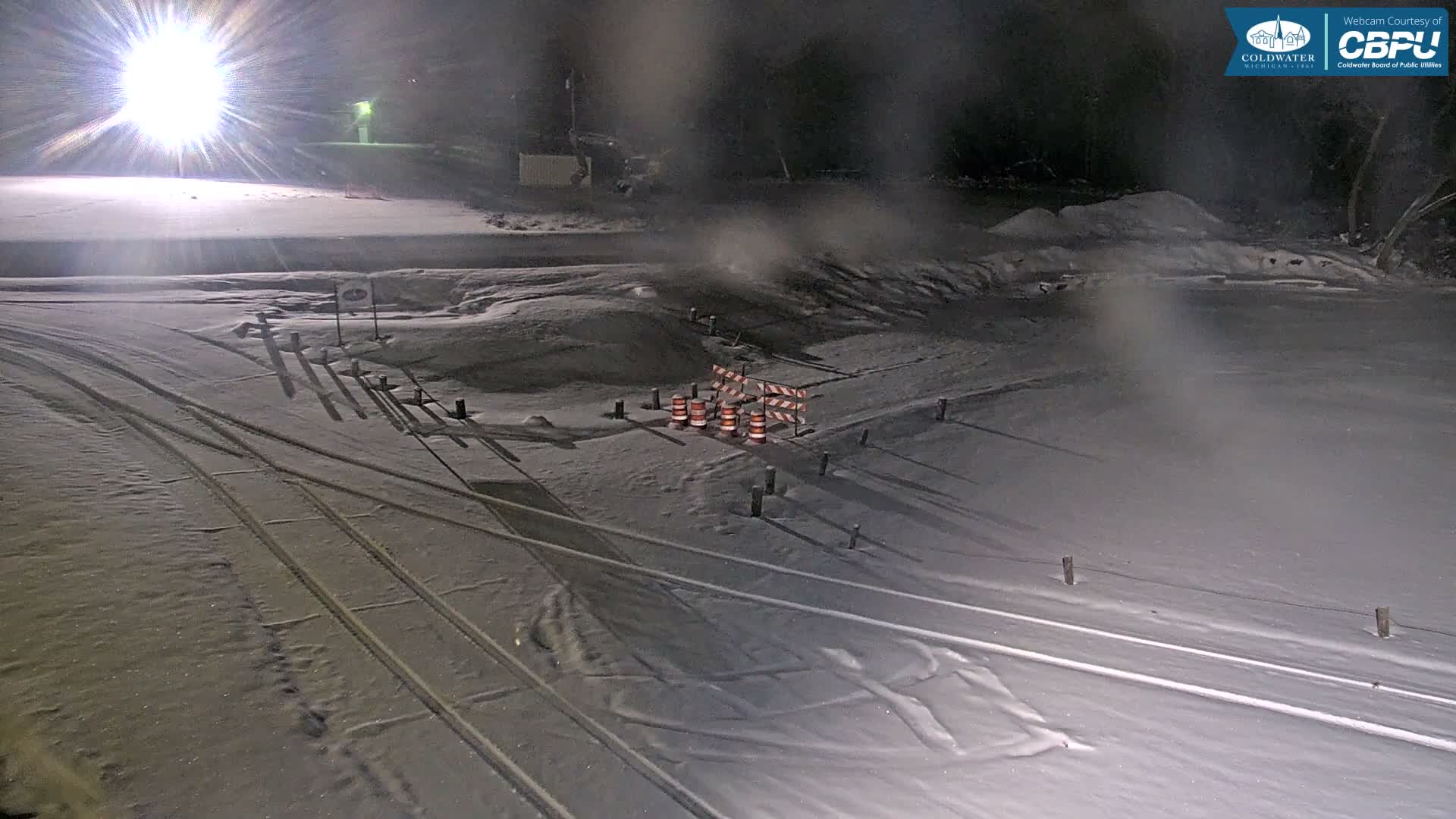 A snow-covered outdoor area at night is illuminated by a bright light source on the left, revealing tracks in the snow, construction barriers, and hazy, wintry conditions.