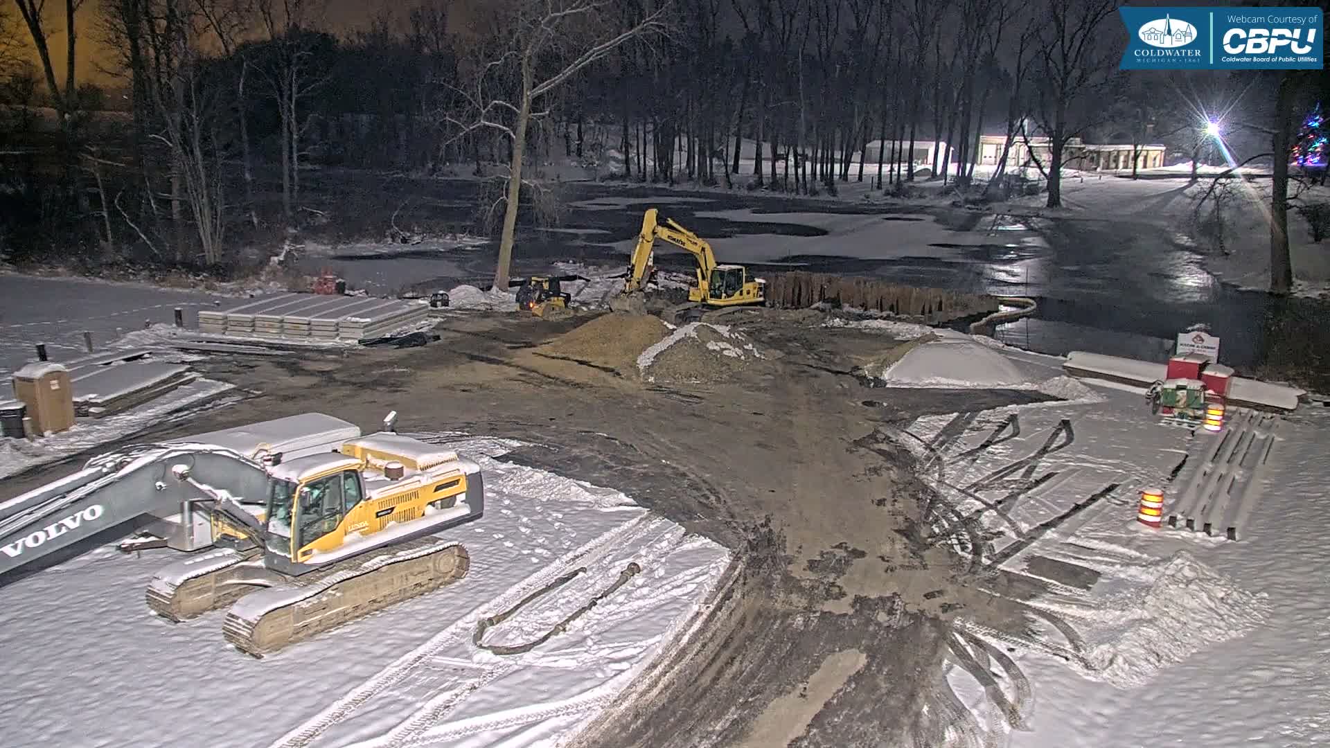 A snowy outdoor construction site at night features heavy machinery, piles of earth, and stacks of materials alongside a partially frozen body of water and bare trees, all under artificial illumination.