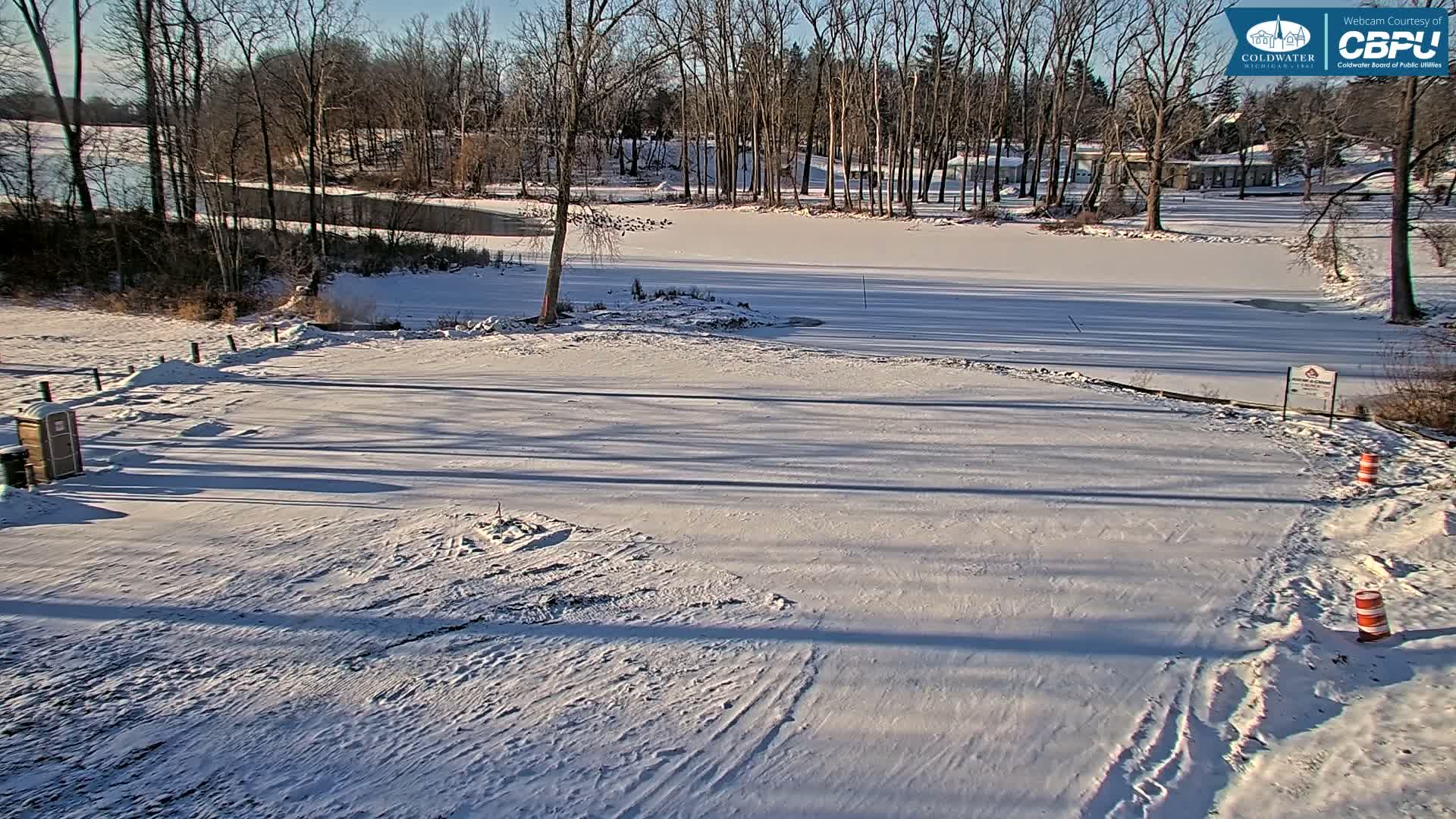 A snowy outdoor construction site at night features heavy machinery, piles of earth, and stacks of materials alongside a partially frozen body of water and bare trees, all under artificial illumination.