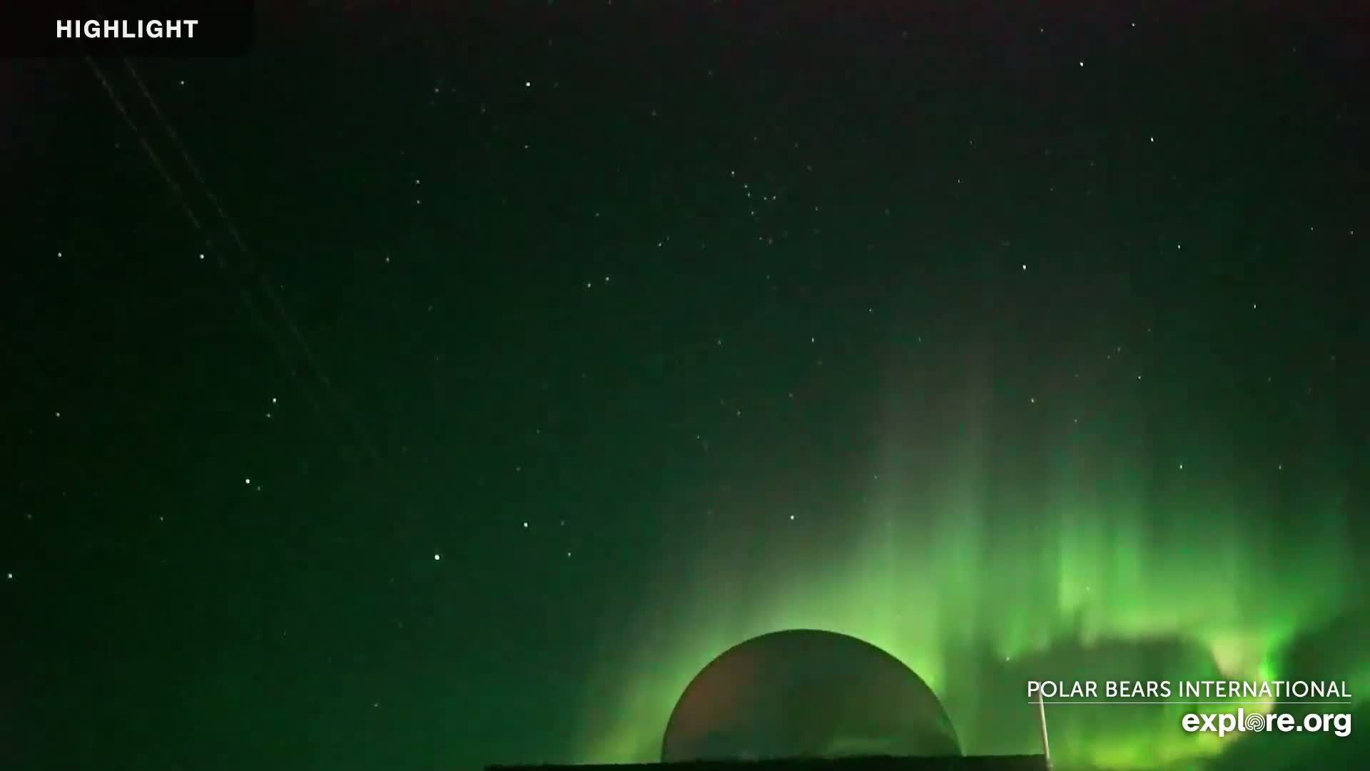 A large, round structure is silhouetted against a night sky filled with stars and a vibrant green aurora borealis.