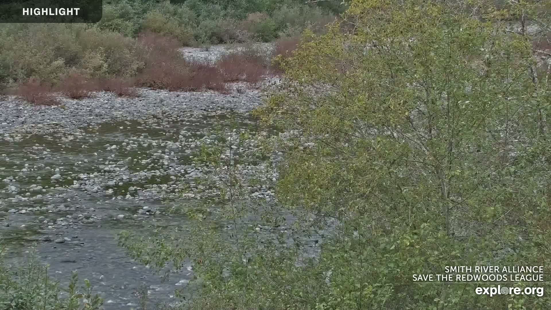 A shallow river with a rocky bed flows through a landscape of green and reddish-brown vegetation under a cloudy sky.