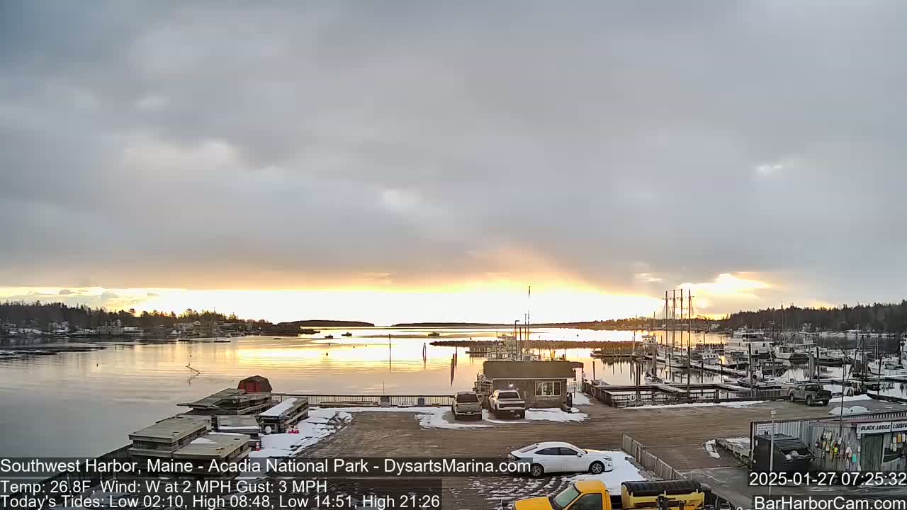 A calm harbor with docked boats at sunrise under a mostly cloudy sky, with a light dusting of snow on the ground.