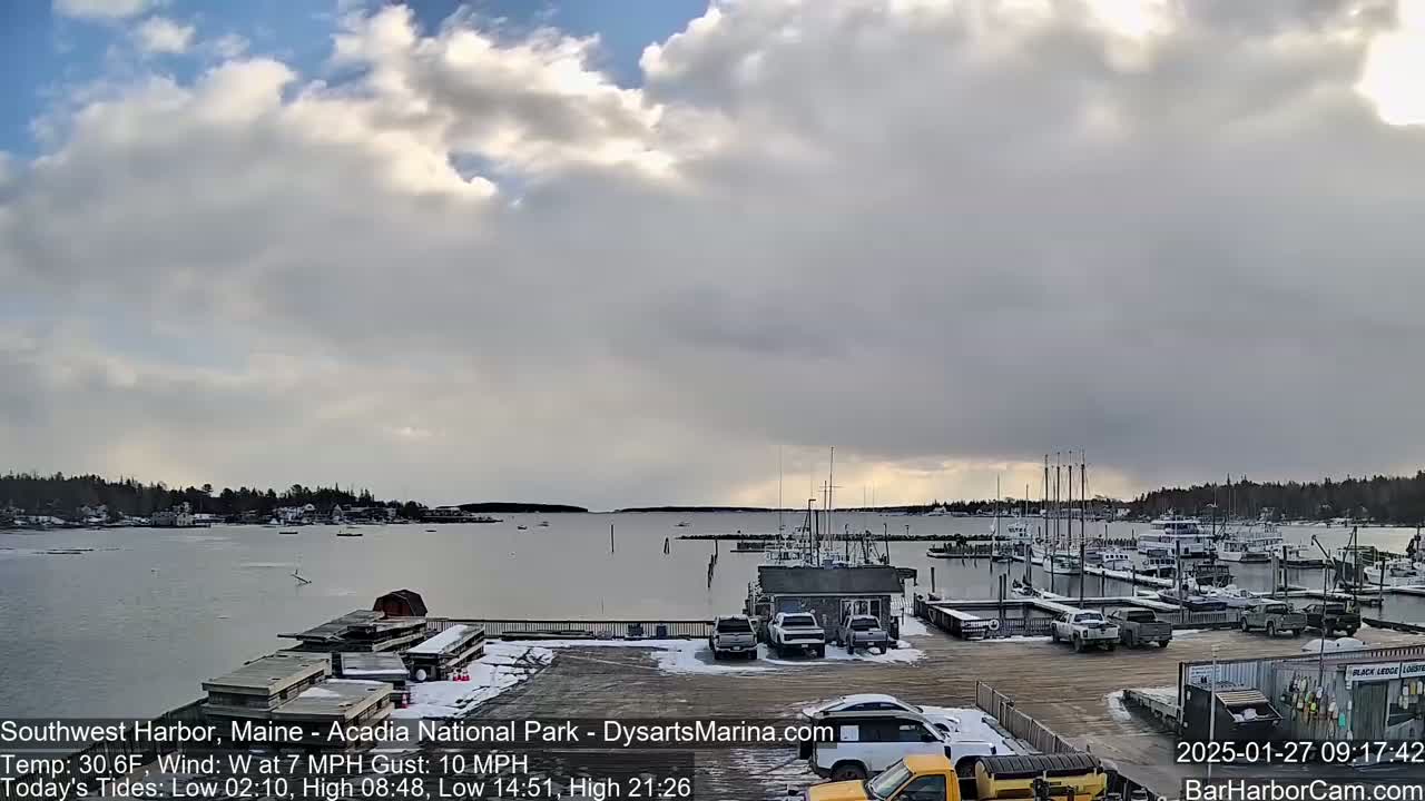 A partly cloudy, cool day shows a harbor with numerous boats docked, a parking lot with several vehicles, and light snow on the ground.