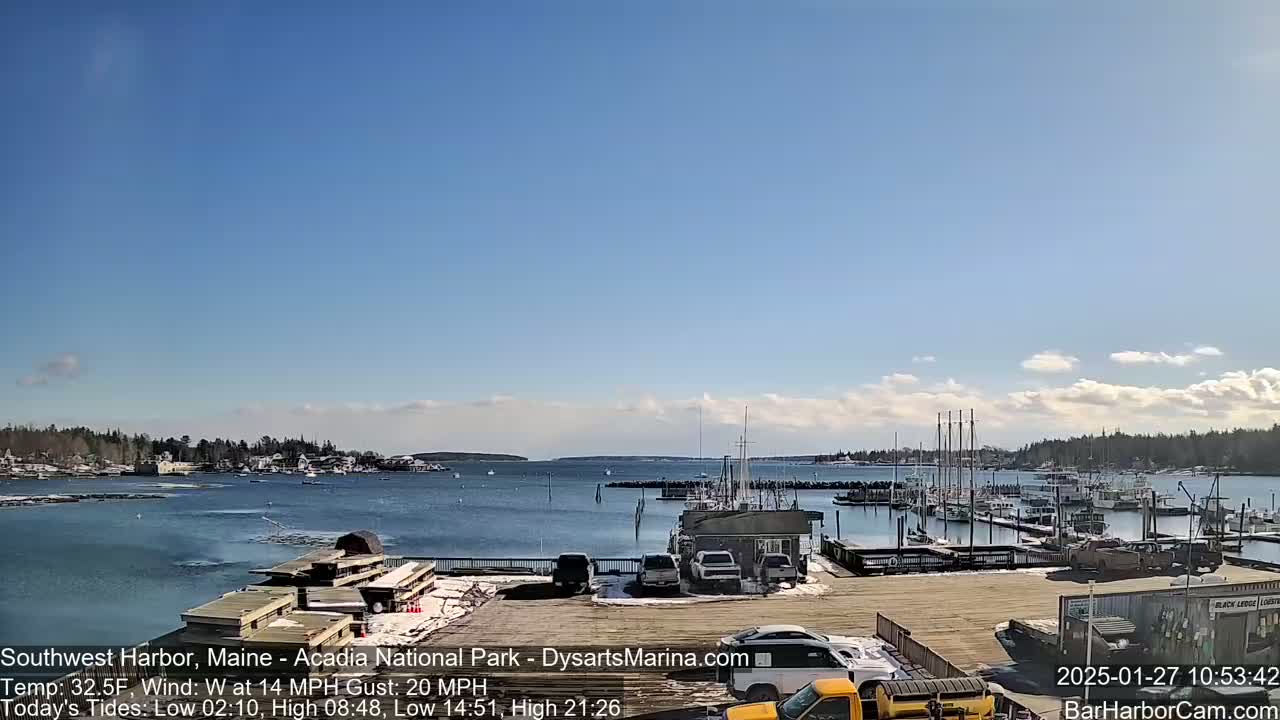 A harbor filled with boats on a sunny day under a mostly clear blue sky, with some light clouds and a light dusting of snow on the ground.