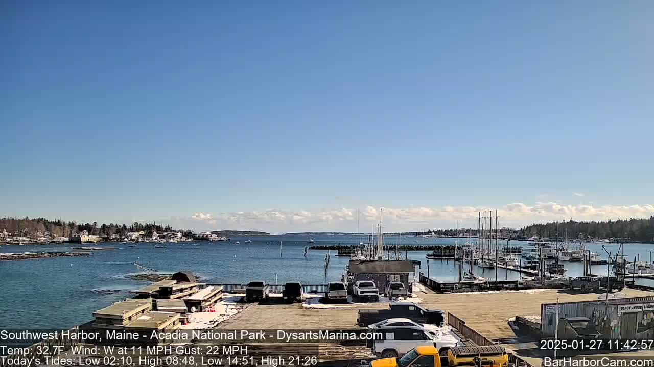 A harbor filled with boats under a mostly clear, bright blue sky with a few clouds, and some snow visible on the ground.