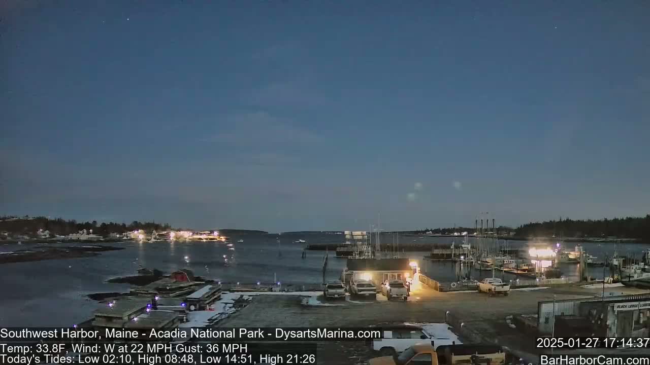 A harbor at twilight, with a calm body of water, some boats docked, and a few vehicles parked near a building, under a mostly clear, dark blue sky.
