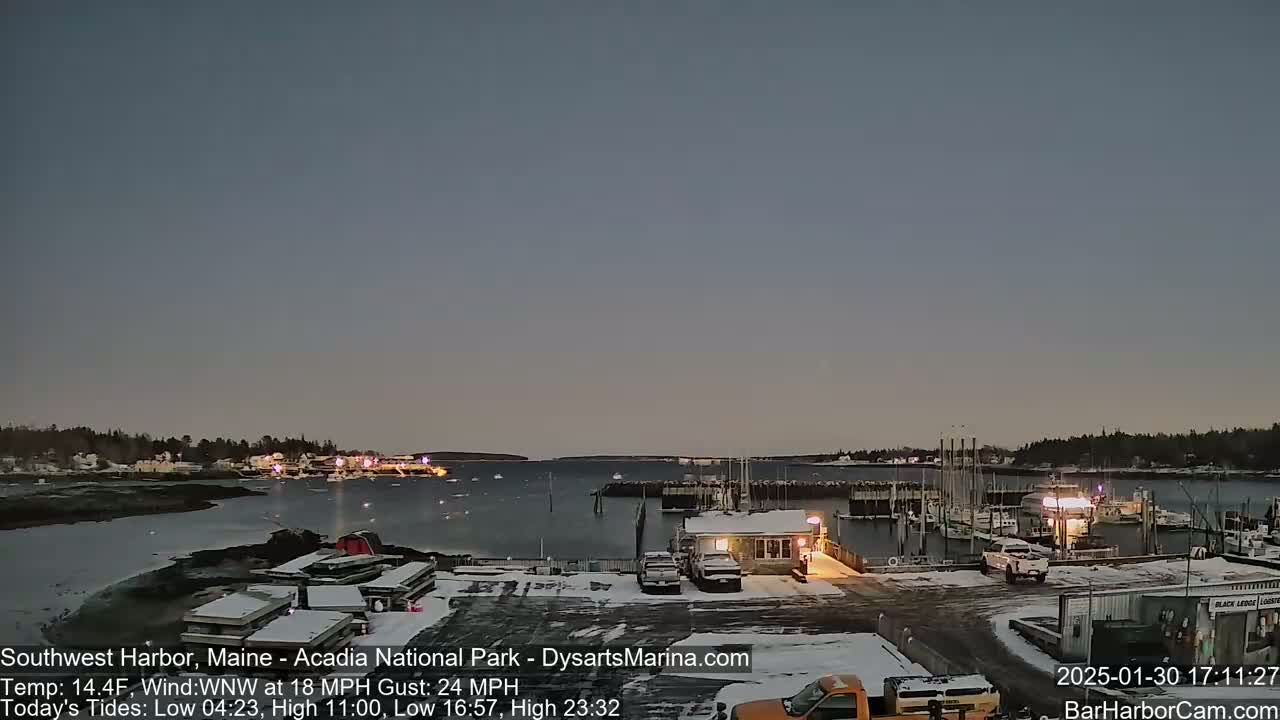 A snow-covered harbor at dusk, with boats moored at a marina and a town visible in the distance under a mostly clear sky.