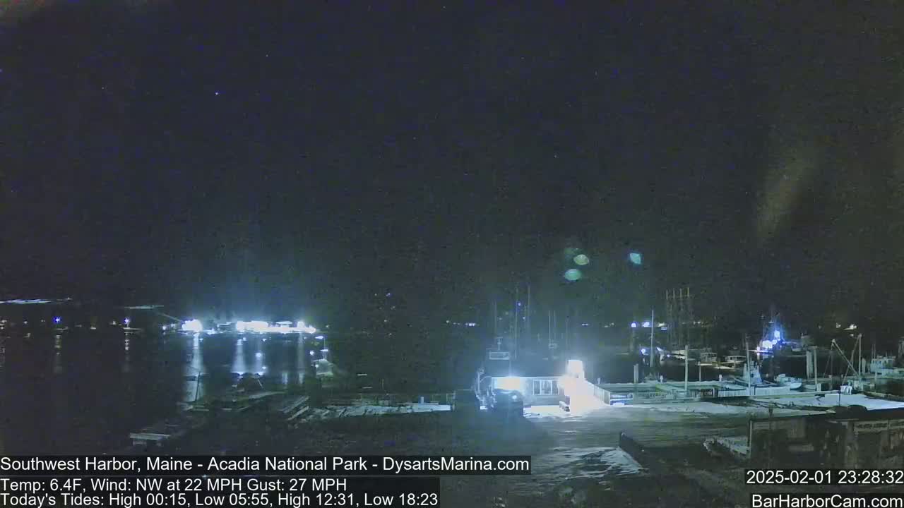A nighttime view of a harbor with boats docked, snow on the ground, and a partly cloudy sky with a northwest wind.