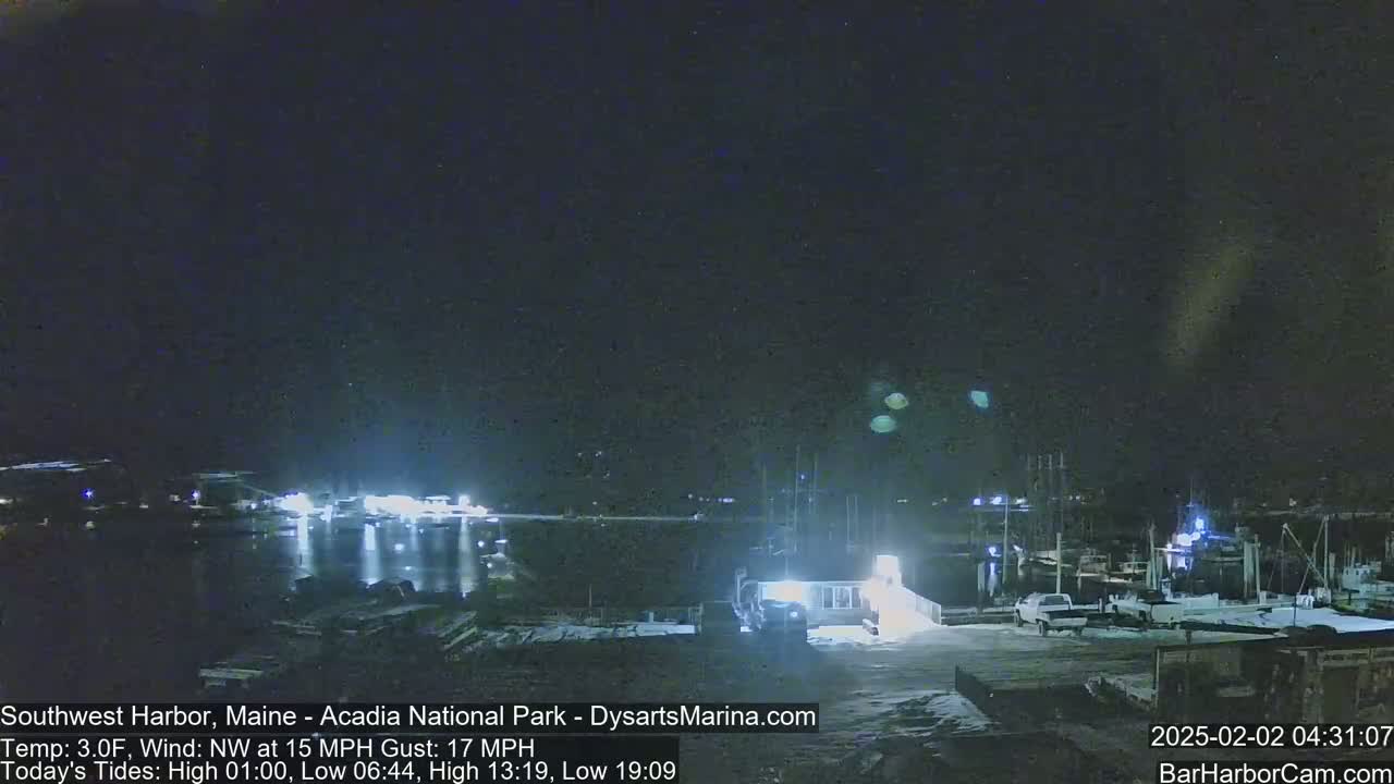 A nighttime harbor scene shows snow-covered ground, docked boats, and a calm body of water reflecting lights under a dark sky.
