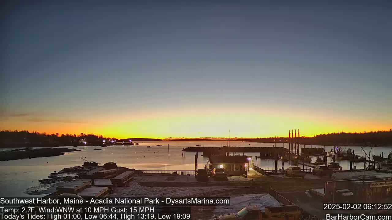 A marina with boats and a small building is shown at sunrise, with a calm body of water under a clear, twilight sky.