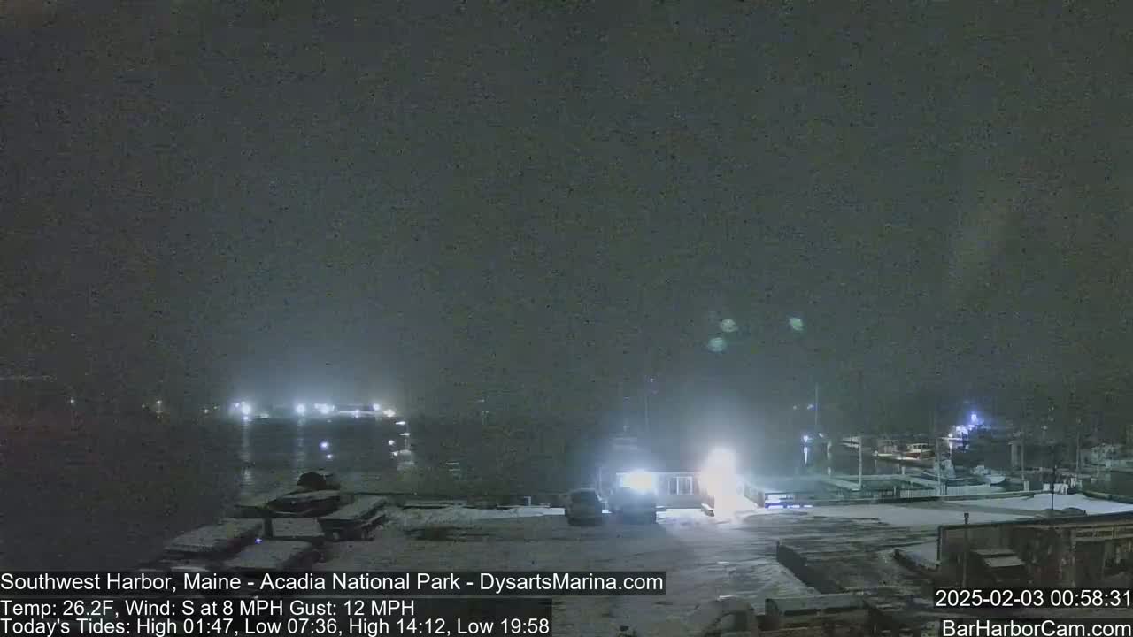 A snow-covered harbor at night, with boats docked and lights illuminating the scene under a dark, overcast sky.