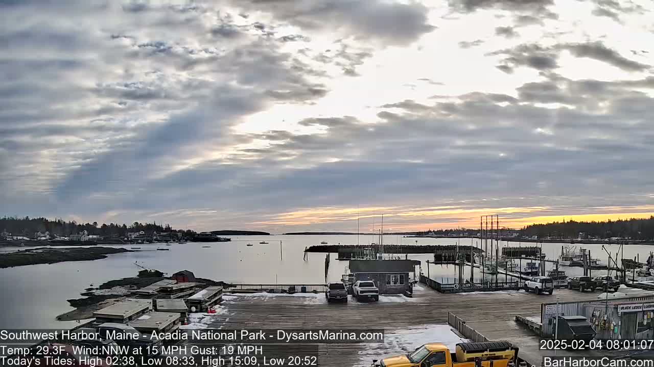 A partly cloudy sunrise over a harbor with docked boats and a few cars parked near a wooden dock, some snow on the ground.