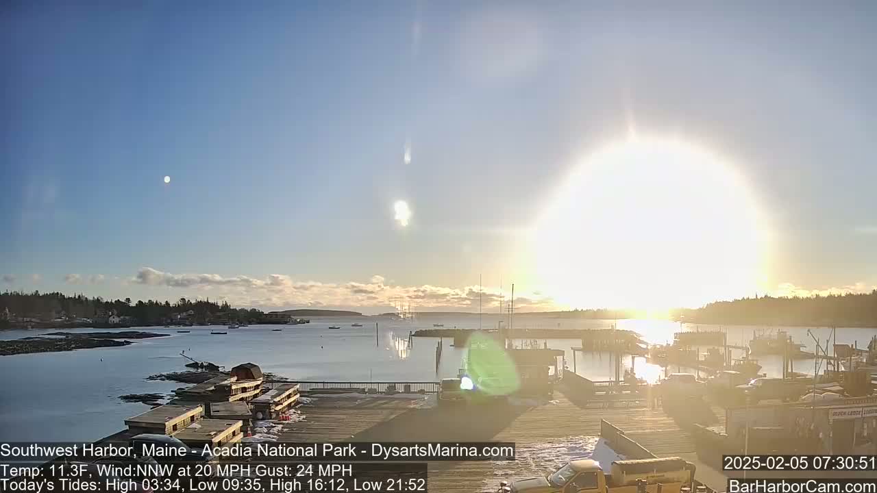 A bright, sunny morning reveals a harbor filled with boats, a town nestled against a shoreline, and a partially snow-covered dock under a mostly clear sky with a light wind.